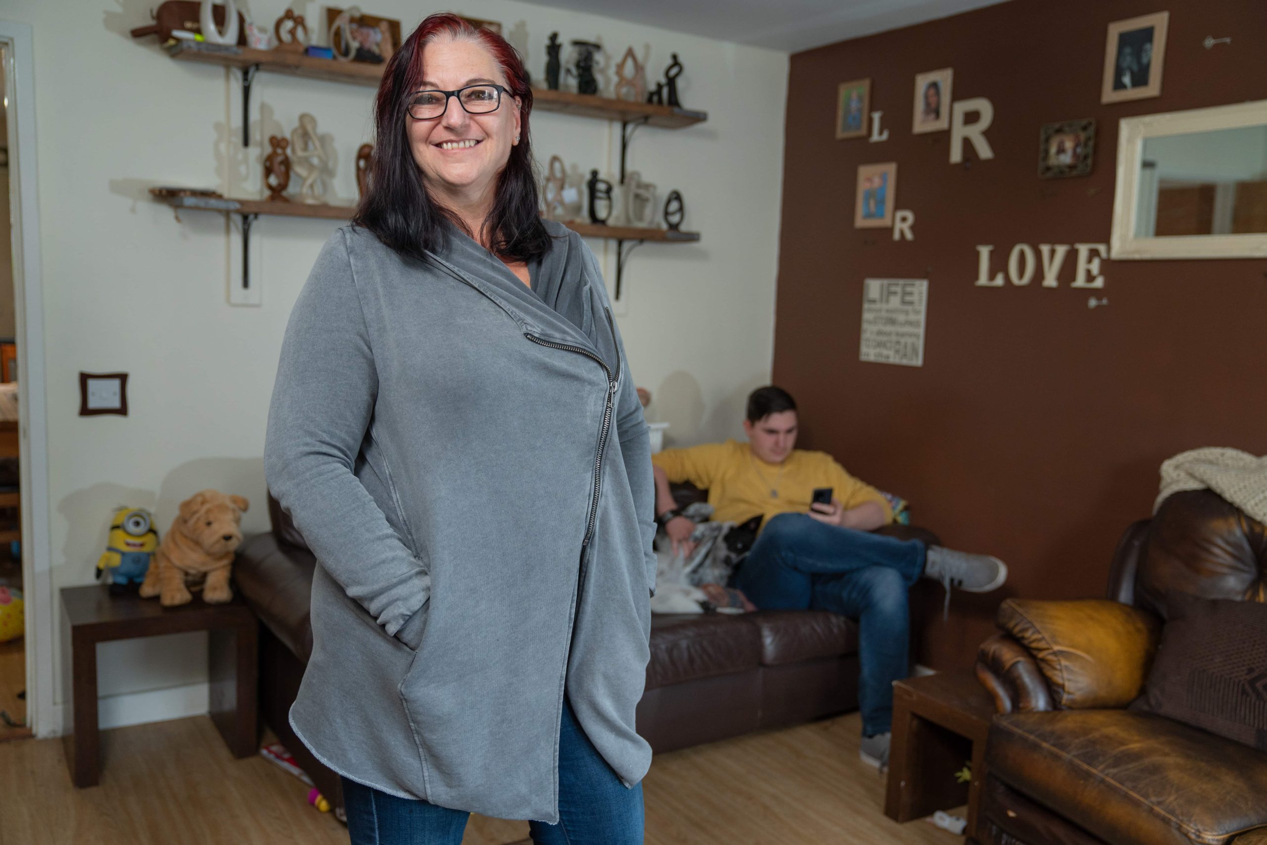 Trivallis Housing Landlord Wales A smiling woman stands in the foreground of a cozy living room, adorned with decorative items on the shelves and wall, sponsored by Trivallis Community Housing. In the background, a young person is focused