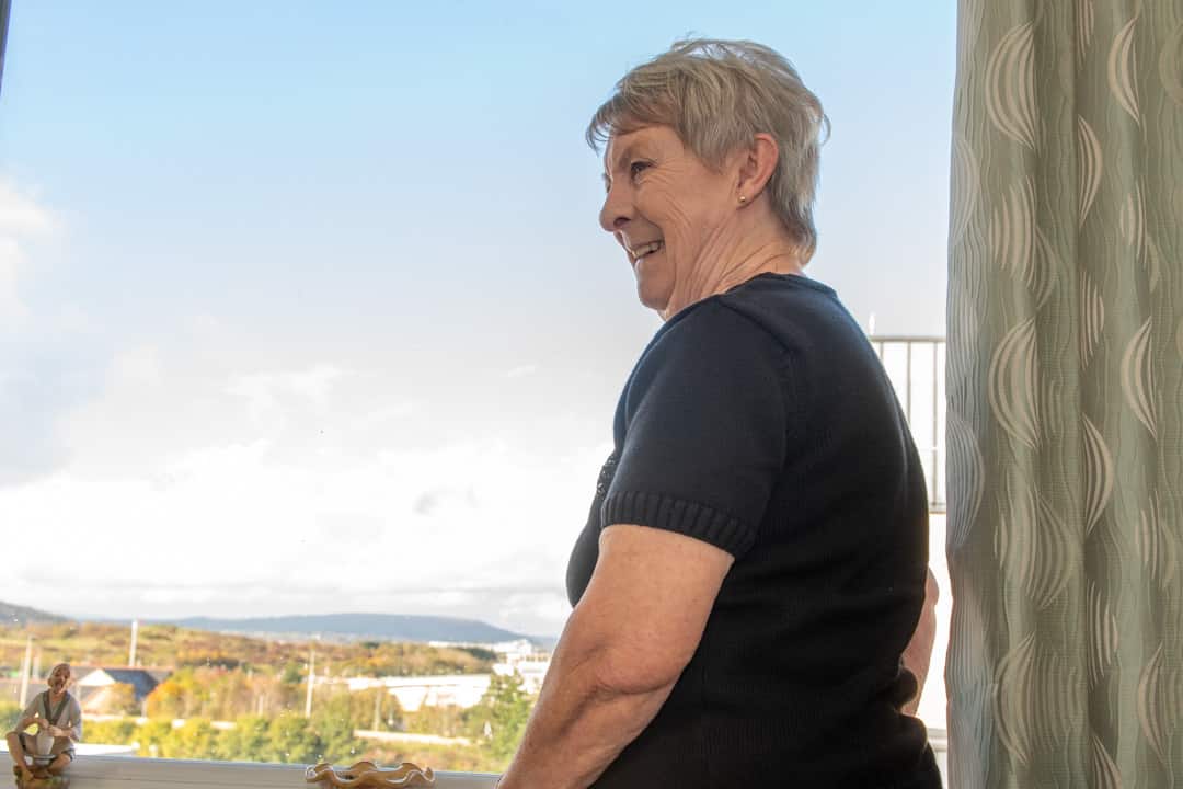 Trivallis Housing Landlord Wales Elderly woman smiling as she looks out a window of a Trivallis Community Housing apartment, basking in natural light, with a serene view in the distance.