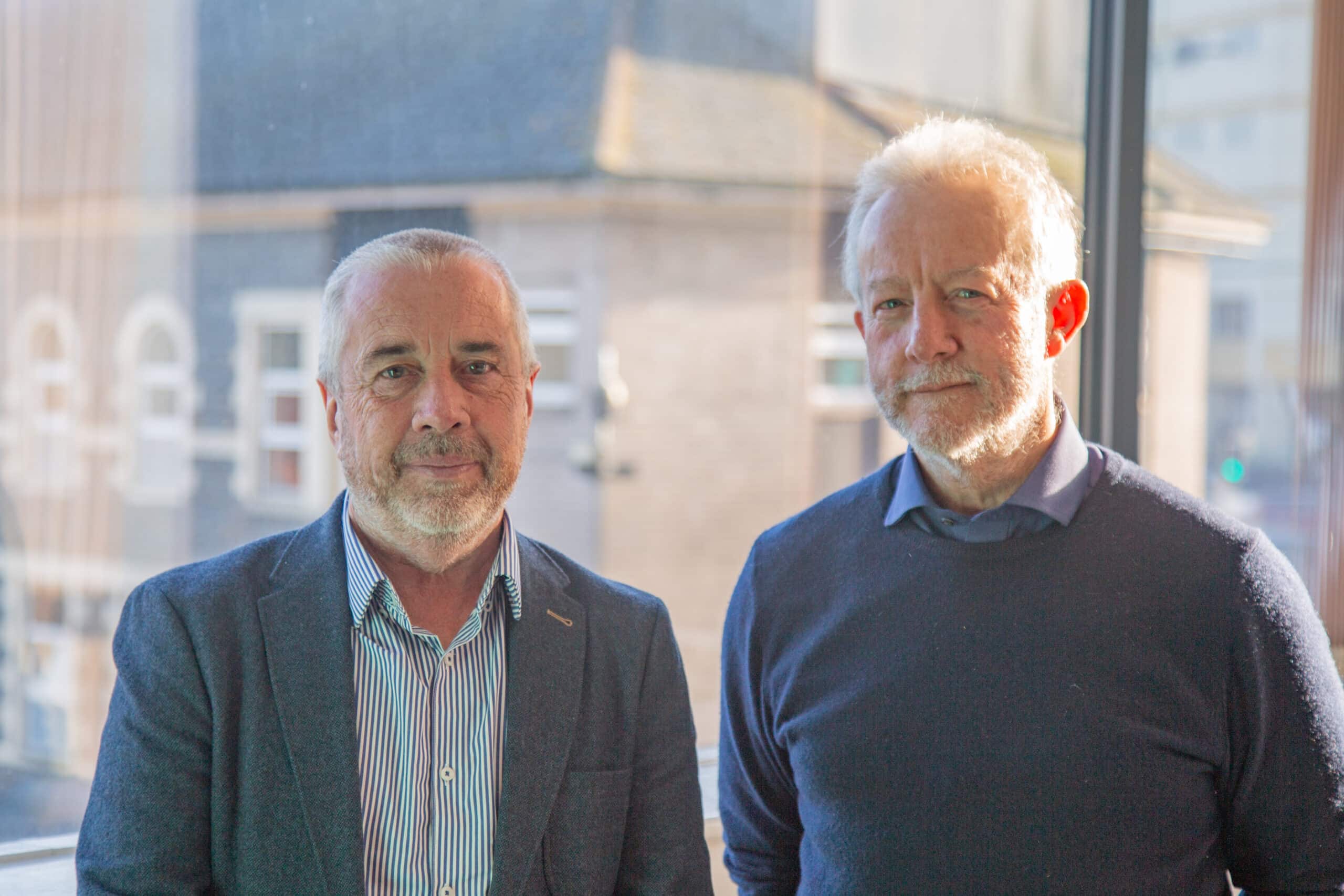 Trivallis Housing Landlord Wales Two middle-aged men standing indoors by a window, with one man slightly in front of the other, both dressed in casual attire with a blurred cityscape of RCT in the background.