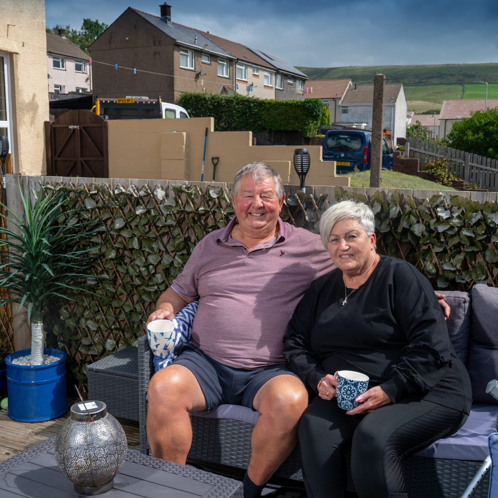 A smiling older couple sitting together on outdoor furniture in a backyard with houses and hills in the background.