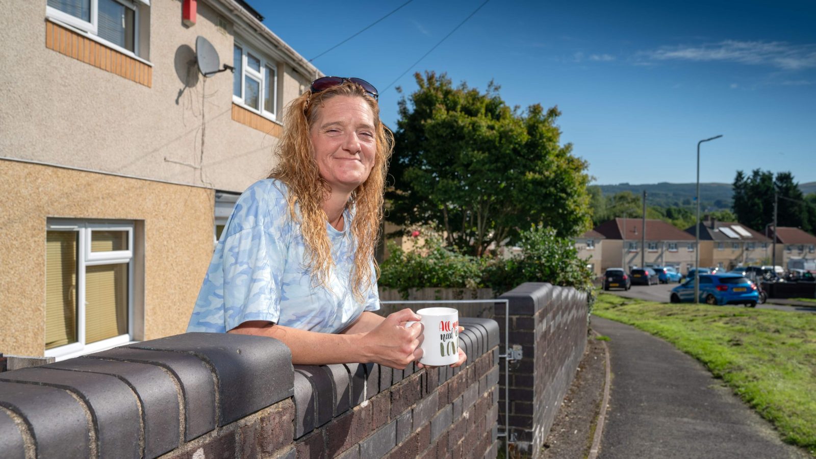 Trivallis Housing Landlord Wales A woman with long hair leaning on a fence, holding a mug, with a Trivallis residential street in the background.