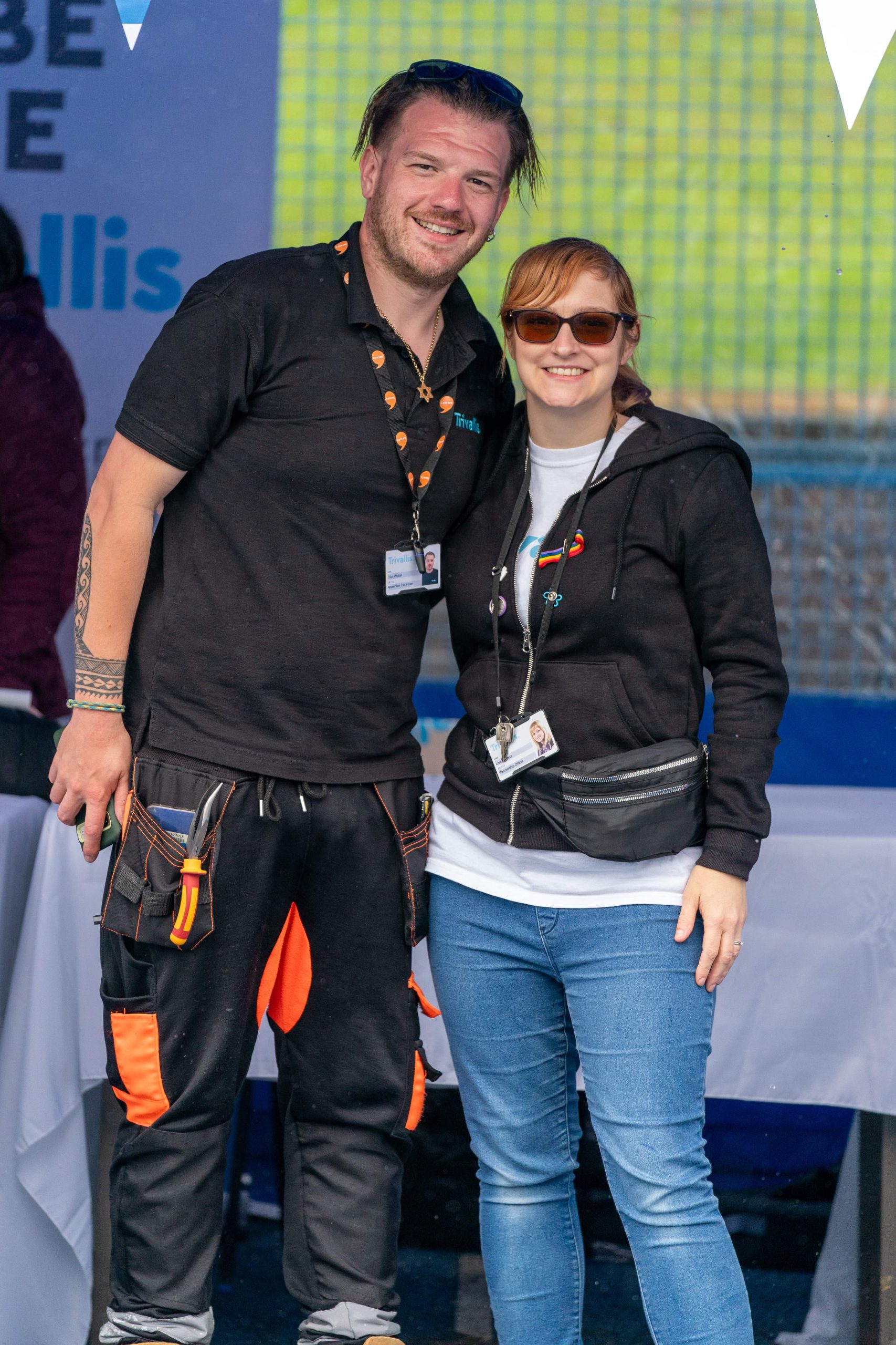 Trivallis Housing Landlord Wales Two individuals, a man and a woman, pose for a photo together, smiling. The man is wearing a black shirt with orange accents and a tool belt, while the woman is in a black hoodie