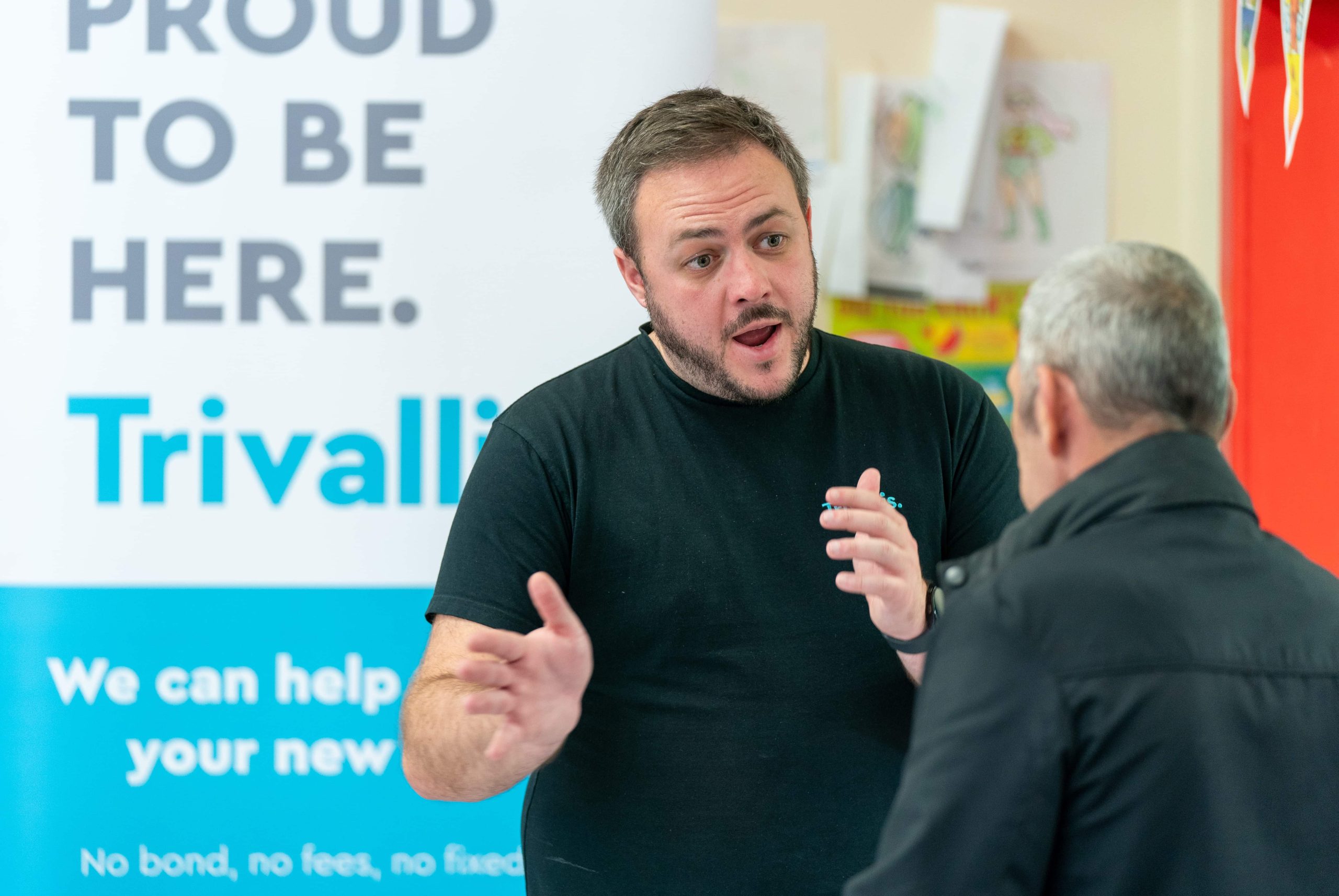 Trivallis Housing Landlord Wales A man gestures while speaking intensely to another person, with a banner reading "proud to be here. Trivallis" visible in the background, highlighting their commitment to housing in RCT.