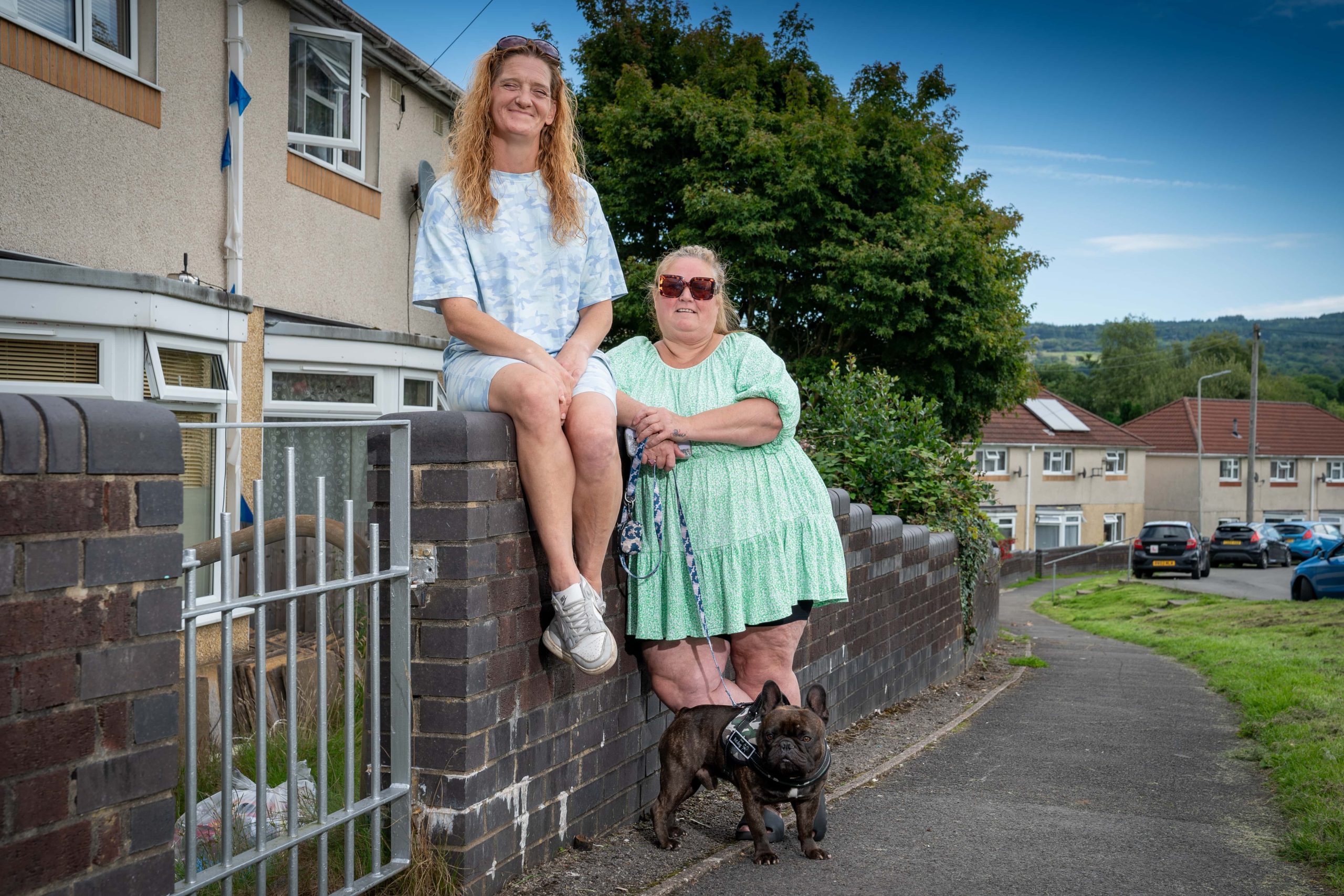 Trivallis Housing Landlord Wales Two women sitting on a low wall outside a row of Trivallis houses, accompanied by a small black dog on a leash.