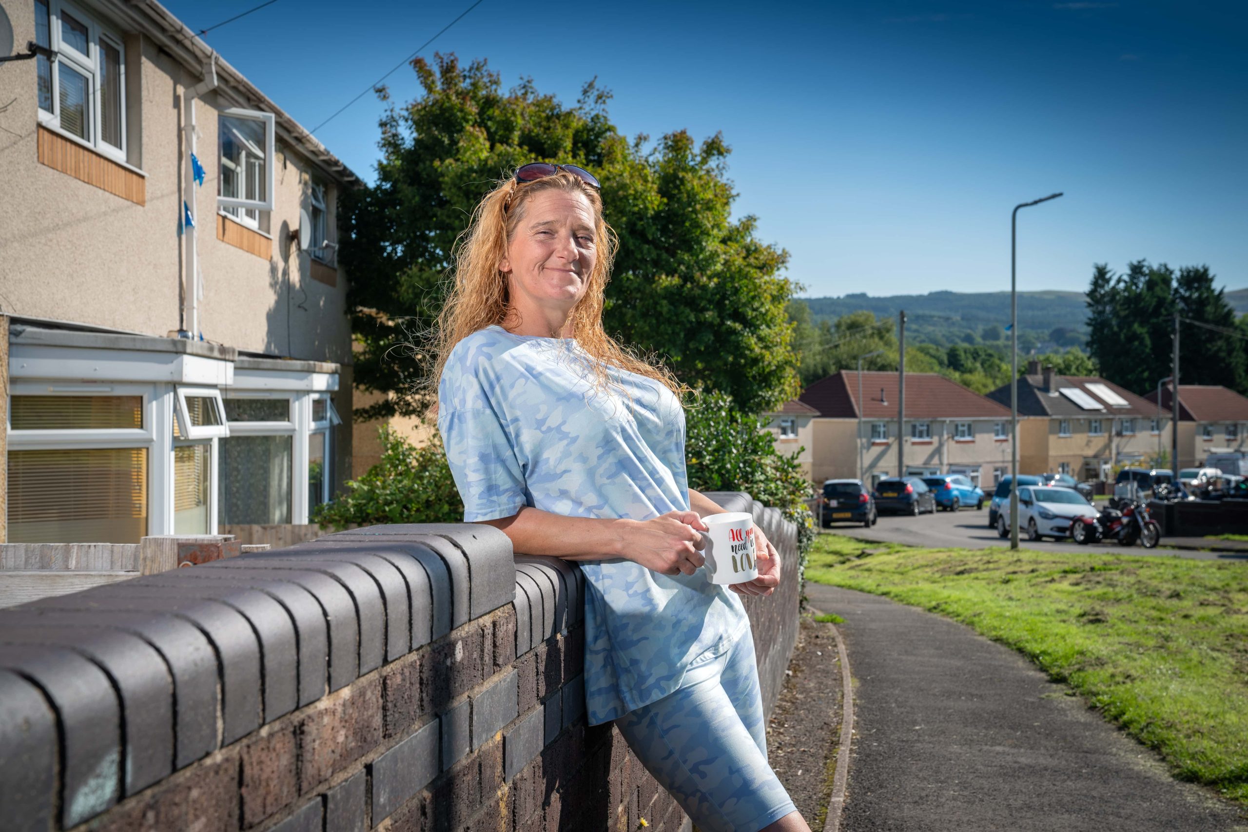 Trivallis Housing Landlord Wales A smiling woman holding a mug stands leaning on a brick wall on a sunny RCT suburban street.