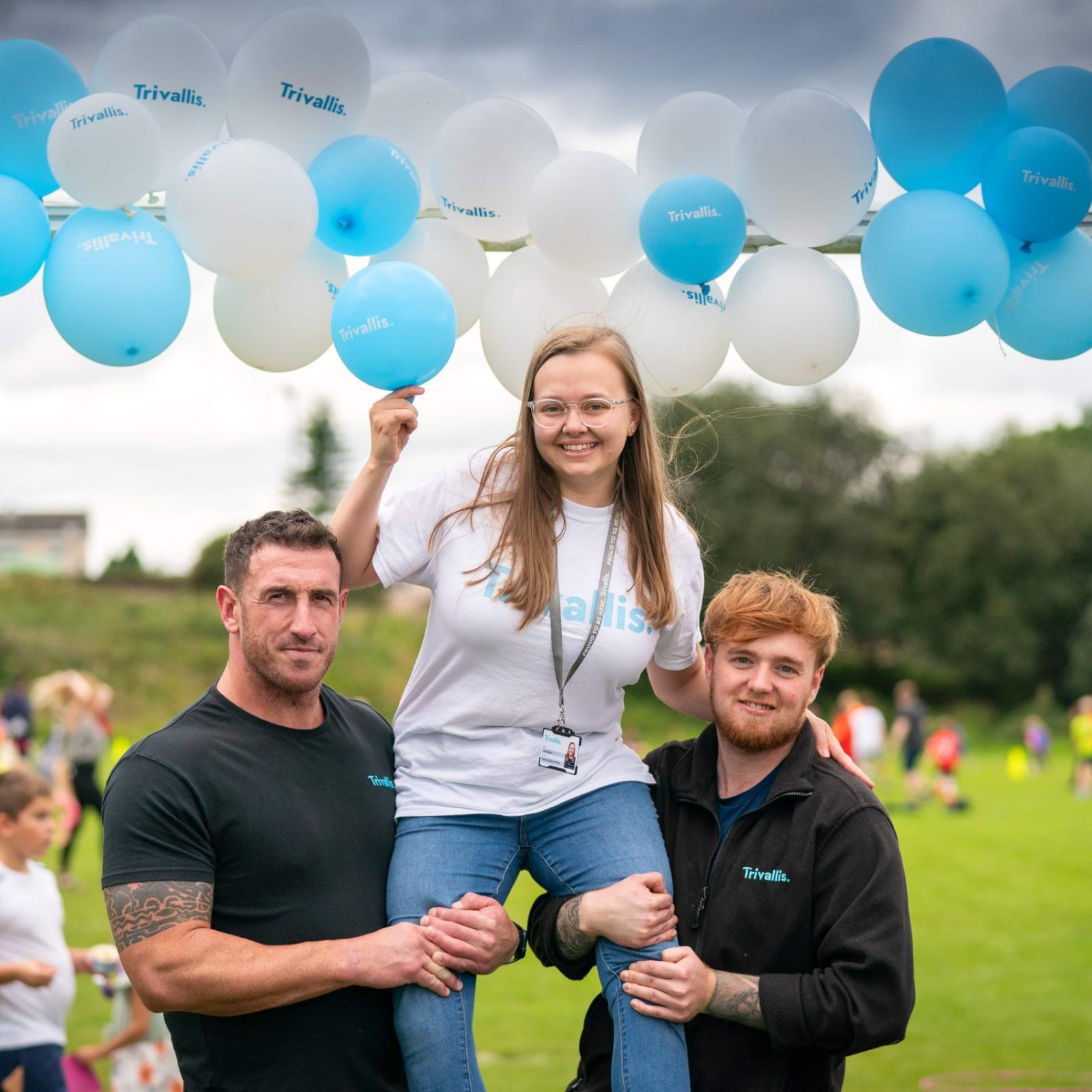 Trivallis Housing Landlord Wales Two individuals lift another person into the air, seemingly lightened by a bunch of blue and white balloons, at an outdoor Trivallis housing event with onlookers in the background.