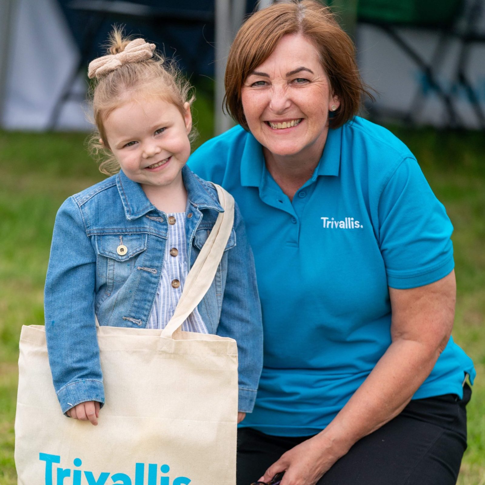 Trivallis Housing Landlord Wales A young girl with a ponytail and a woman in a blue Trivallis shirt are smiling at the camera while kneeling on grass, holding a tote bag with the RCT housing logo.