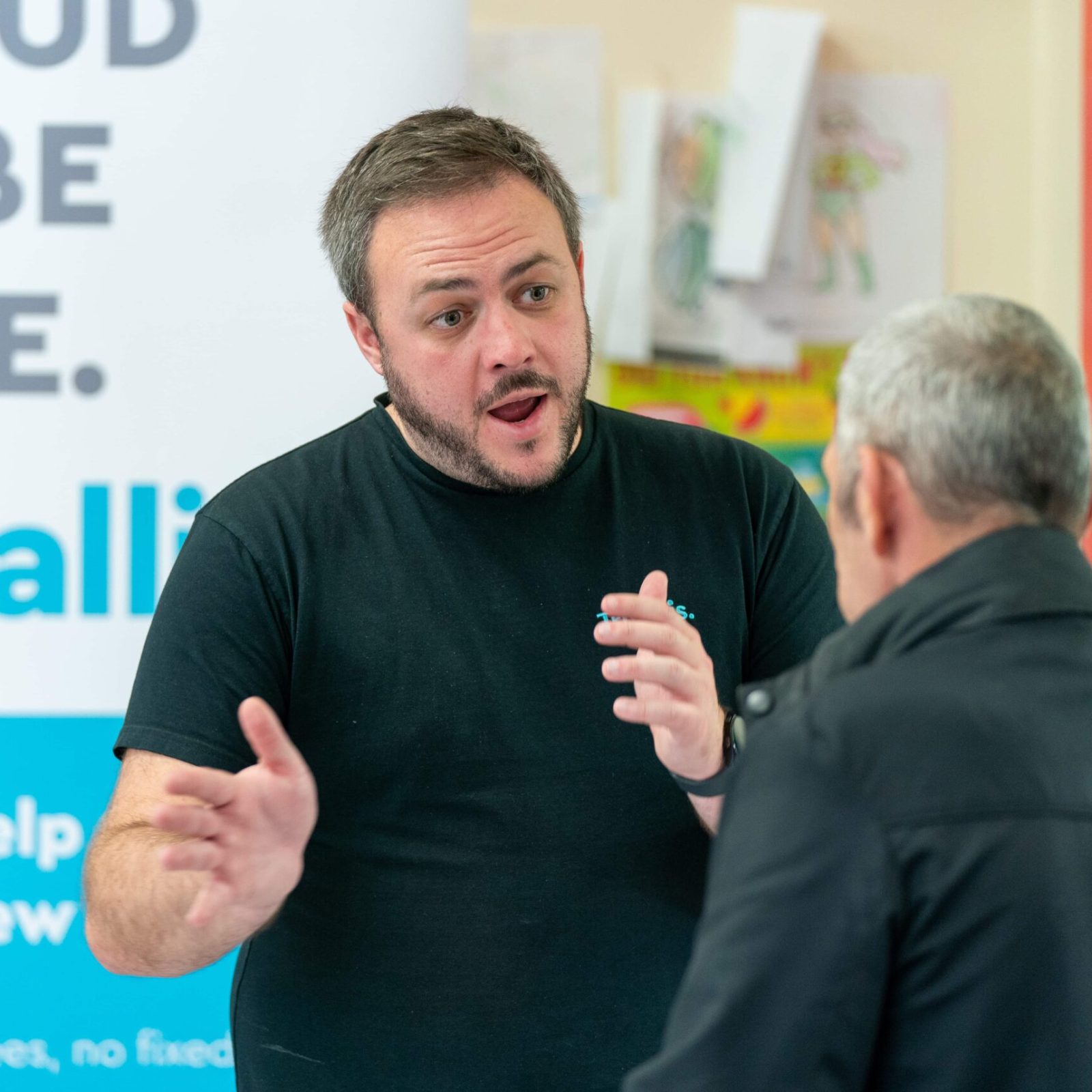 Trivallis Housing Landlord Wales Two men engaged in a conversation, with one man speaking and gesturing with his hands while the other listens, in front of a sign that says 