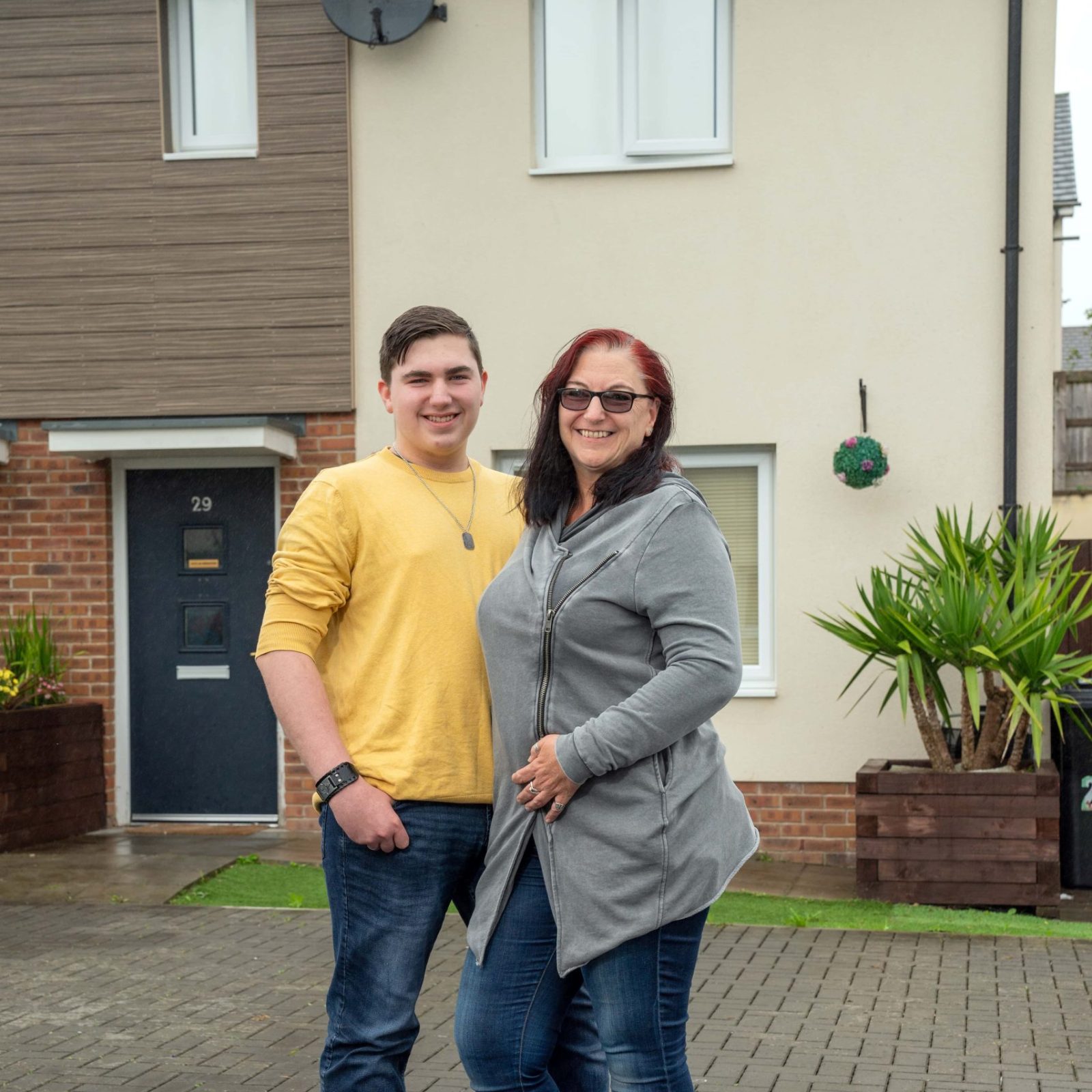 Trivallis Housing Landlord Wales A woman and a younger male companion, possibly her son, are standing together in front of a Trivallis housing property with the visible house number 29 on a cloudy day. Both are smiling and