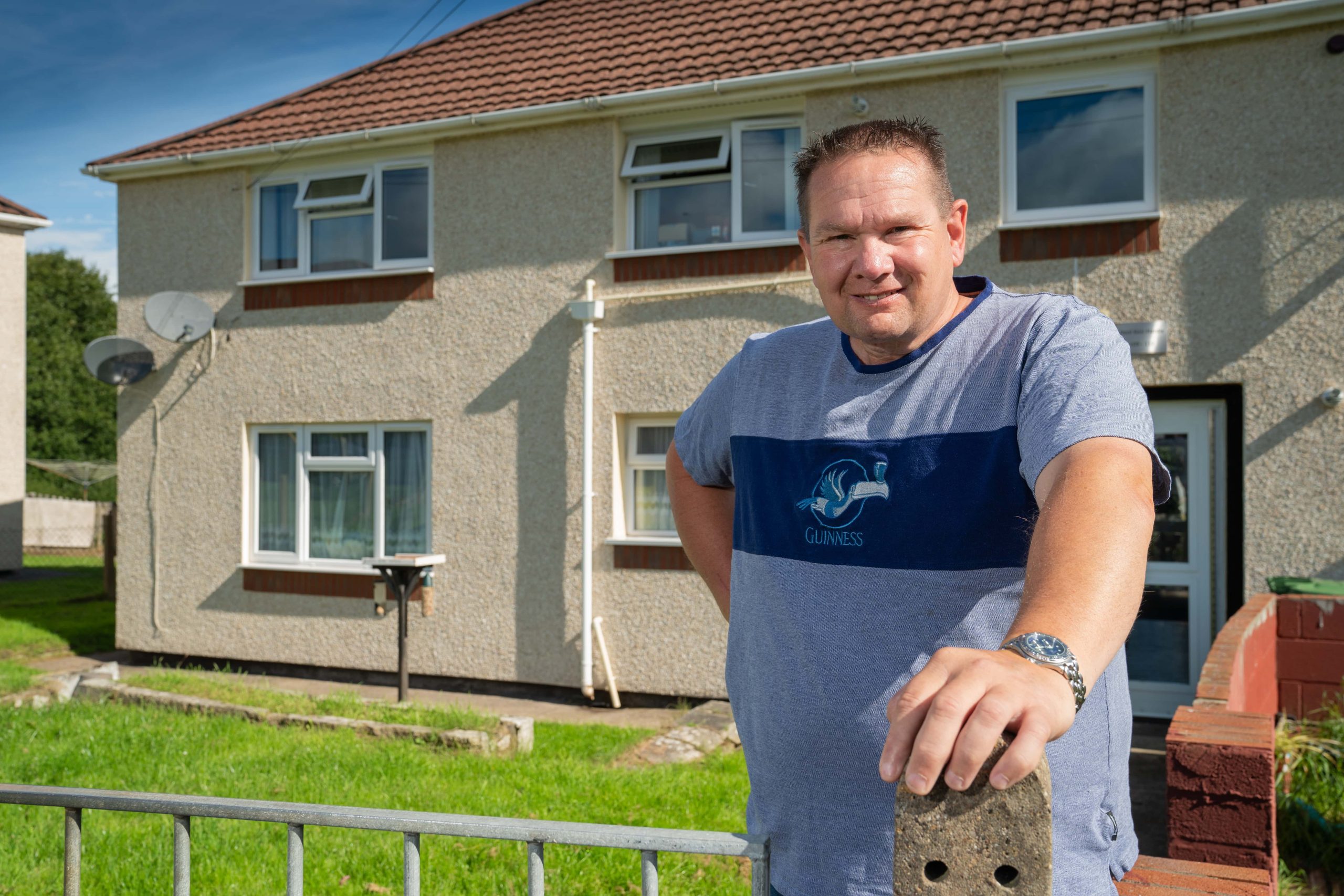 Trivallis Housing Landlord Wales A man stands in front of a two-story Trivallis house with a satellite dish on a sunny day, leaning on a metal fence with green grass in the background.