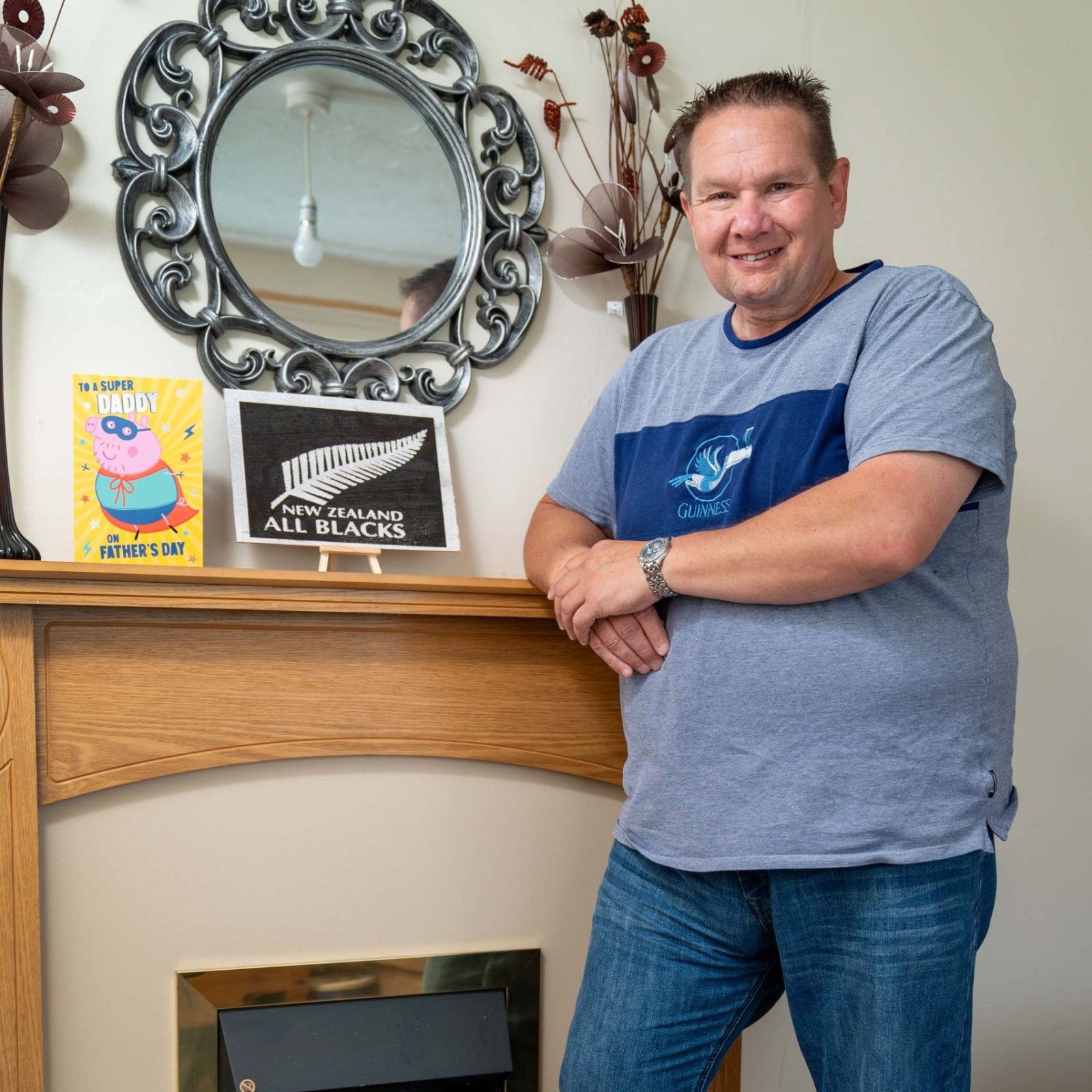 Trivallis Housing Landlord Wales A man stands smiling beside a fireplace mantle adorned with decorative items, a mirror, and a few books in Trivallis housing, RCT.