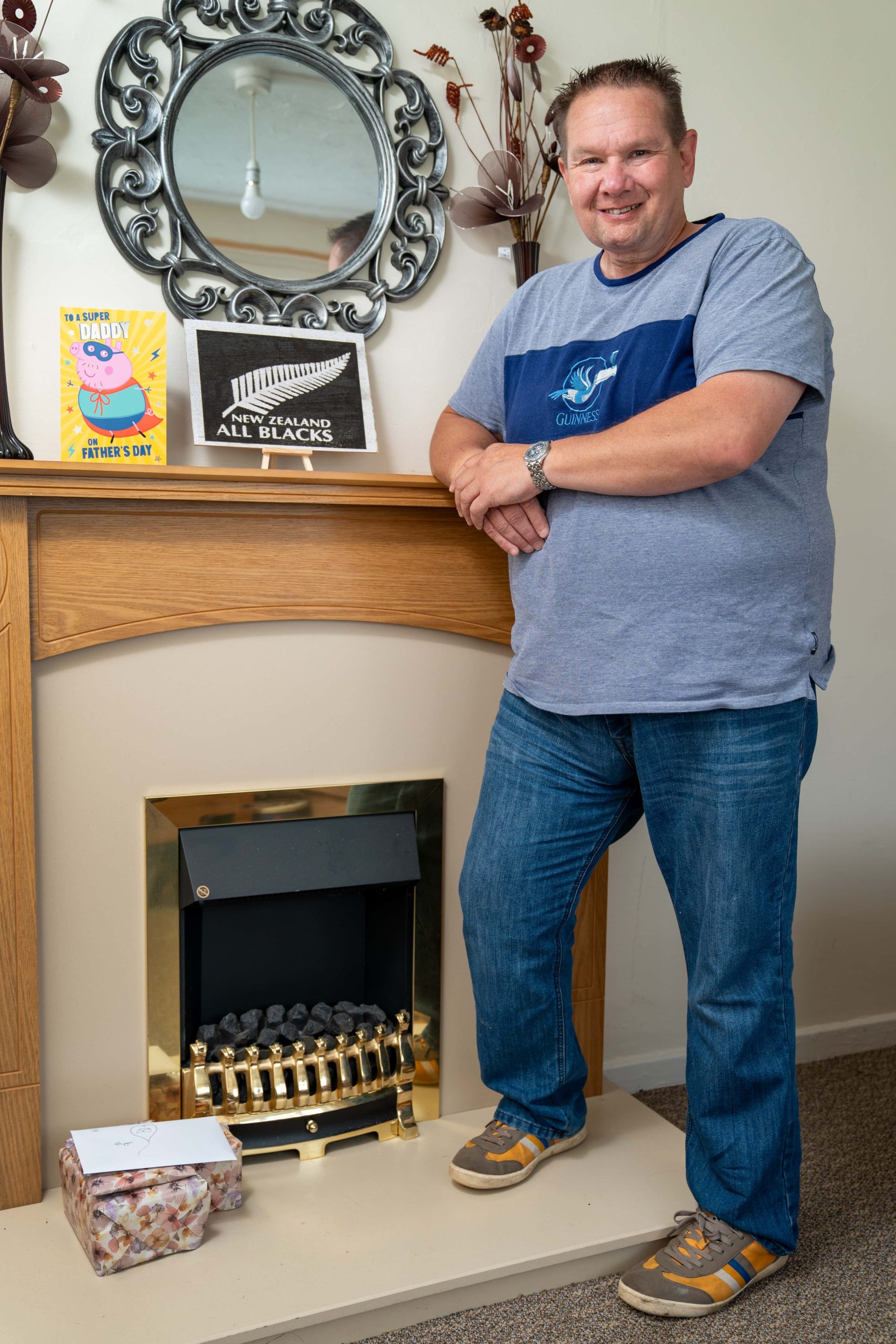 Trivallis Housing Landlord Wales A man stands smiling beside a fireplace mantle adorned with decorative items, a mirror, and a few books in Trivallis housing, RCT.