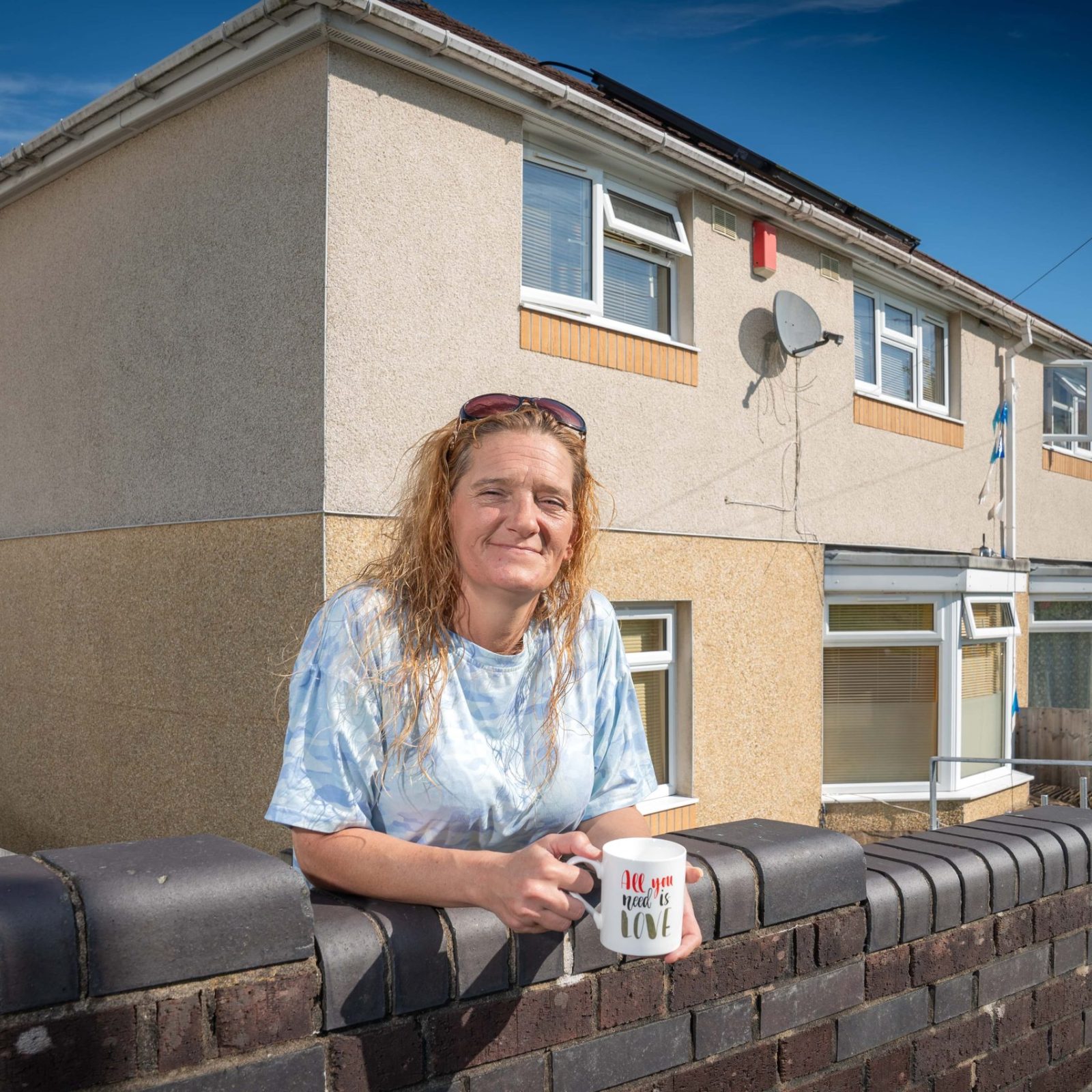 Trivallis Housing Landlord Wales A woman leaning on a brick fence in front of a Trivallis residential building, holding a mug and smiling at the camera.