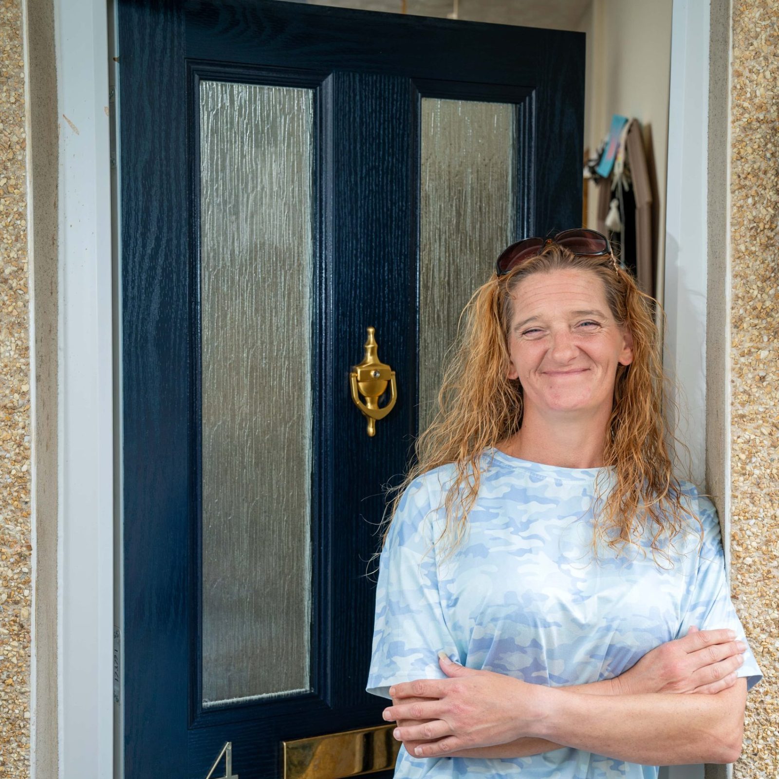 Trivallis Housing Landlord Wales A woman with long hair, wearing a blue and white patterned outfit, stands smiling with her arms crossed in front of a dark blue door marked with the number 4, emblematic of Triv