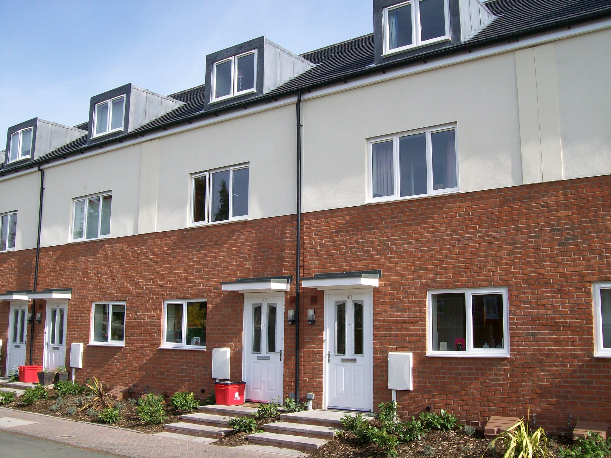 Newly constructed terraced houses with evident brickwork, white doors, and dormer windows under a clear sky.