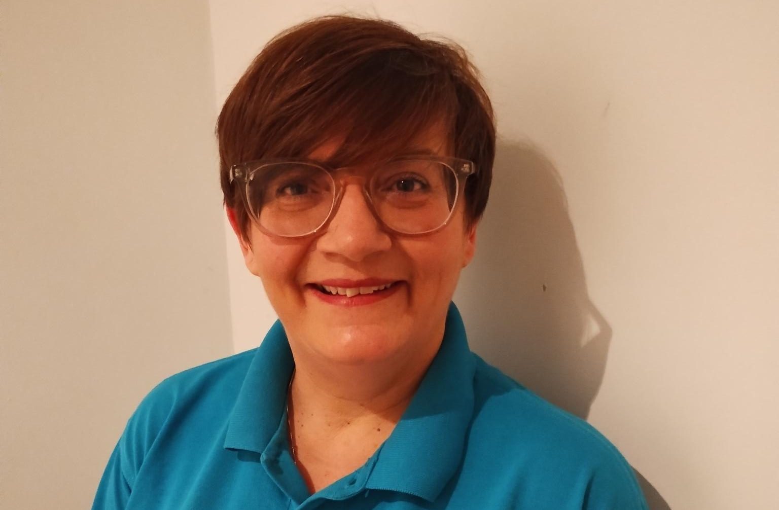 A woman with short brown hair wearing glasses and a blue polo shirt with the logo "trivallis" smiles warmly against a plain white background.