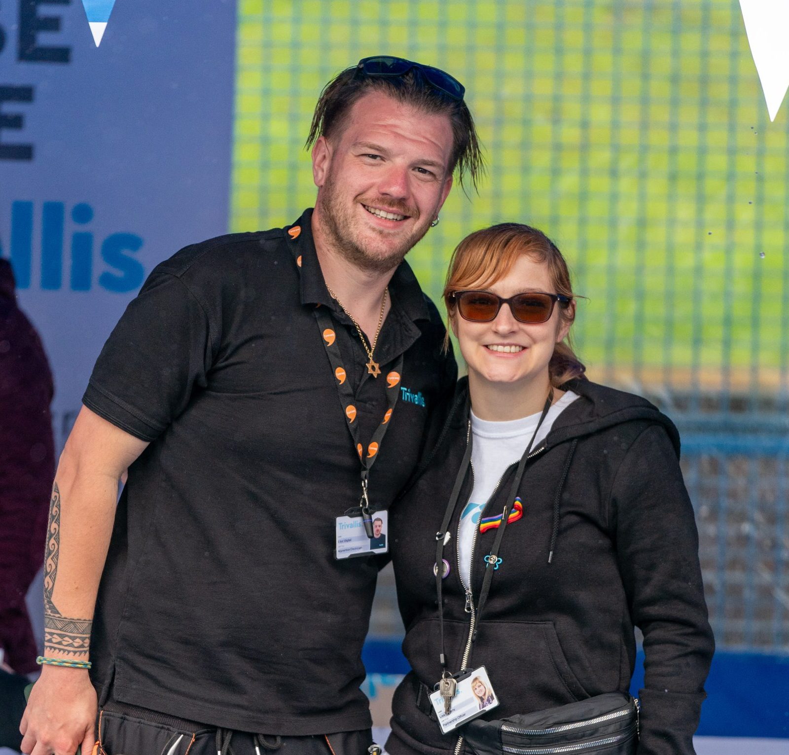 Two people, a man and a woman, smiling at the camera at an outdoor event. both wearing black shirts and jeans, and the man has a tool belt. they're wearing lanyards with ids.