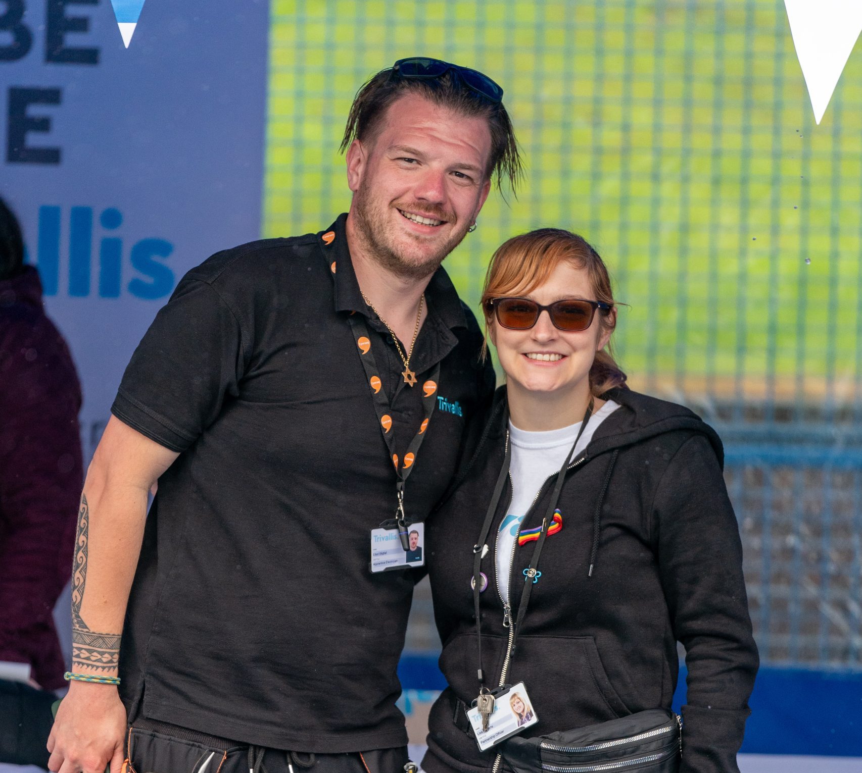 Two people, a man and a woman, smiling at the camera at an outdoor event. both wearing black shirts and jeans, and the man has a tool belt. they're wearing lanyards with ids.