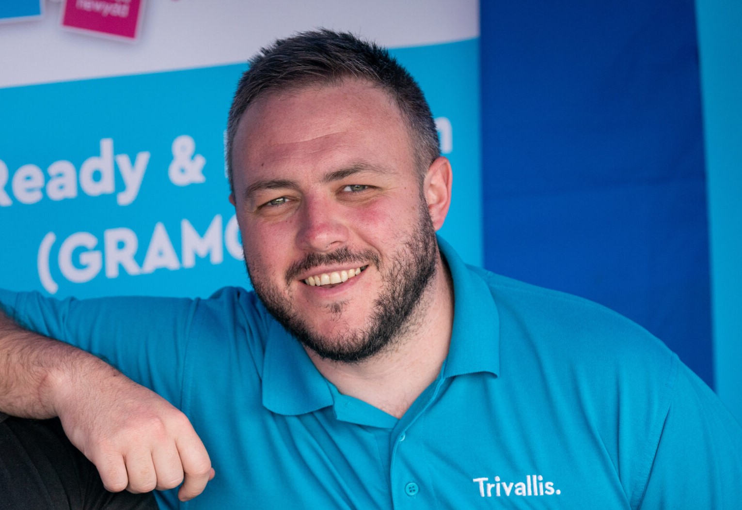 Man in a blue polo shirt with "trivallis" logo smiles at the camera, seated against a blue background with text banners.