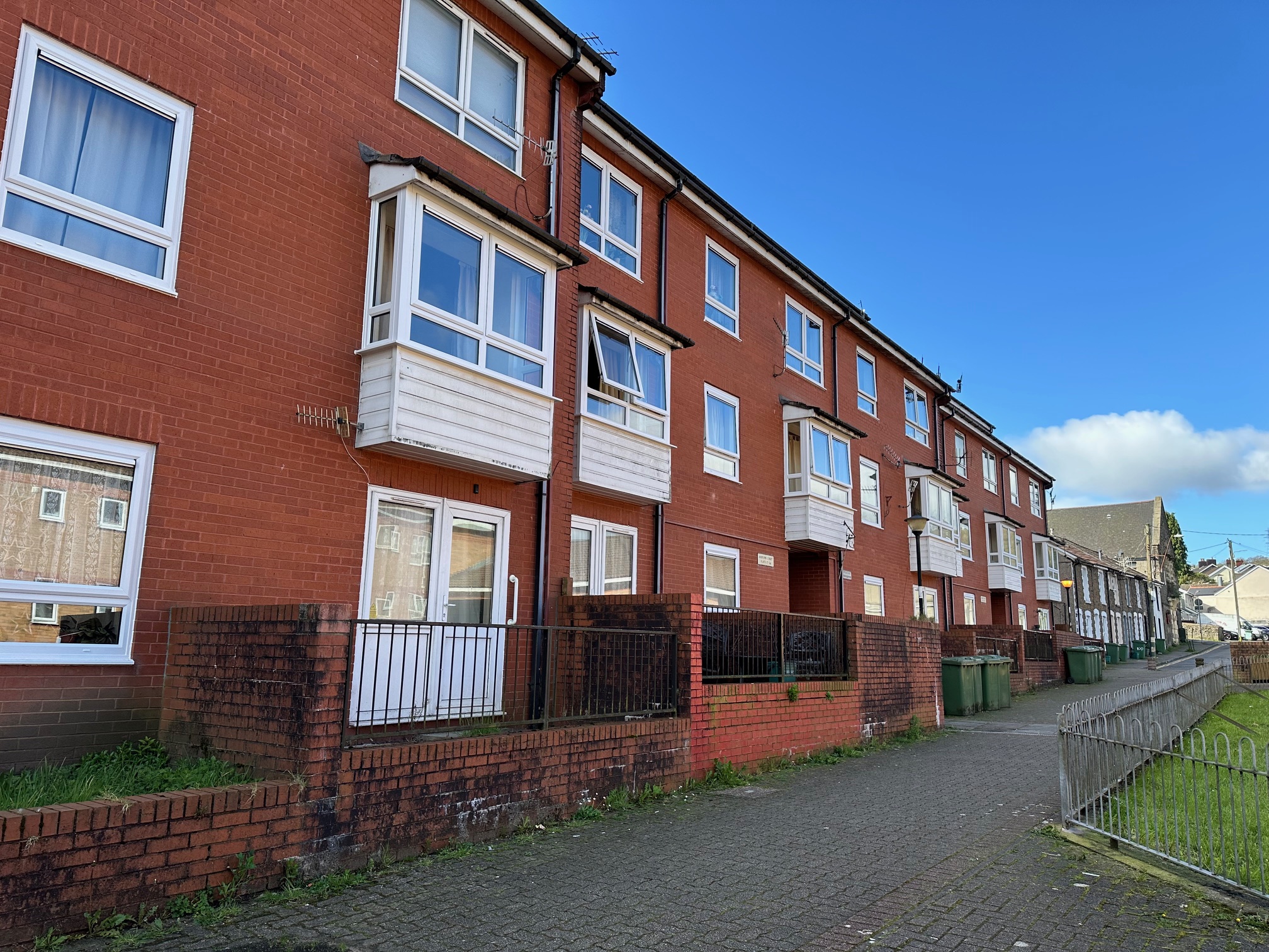 Trivallis Housing Landlord Wales A row of red brick, three-story terraced houses with white-framed windows and balconies. The buildings have small fenced front areas next to a paved walkway. Green garbage bins are visible beside the houses. The sky is clear and blue.