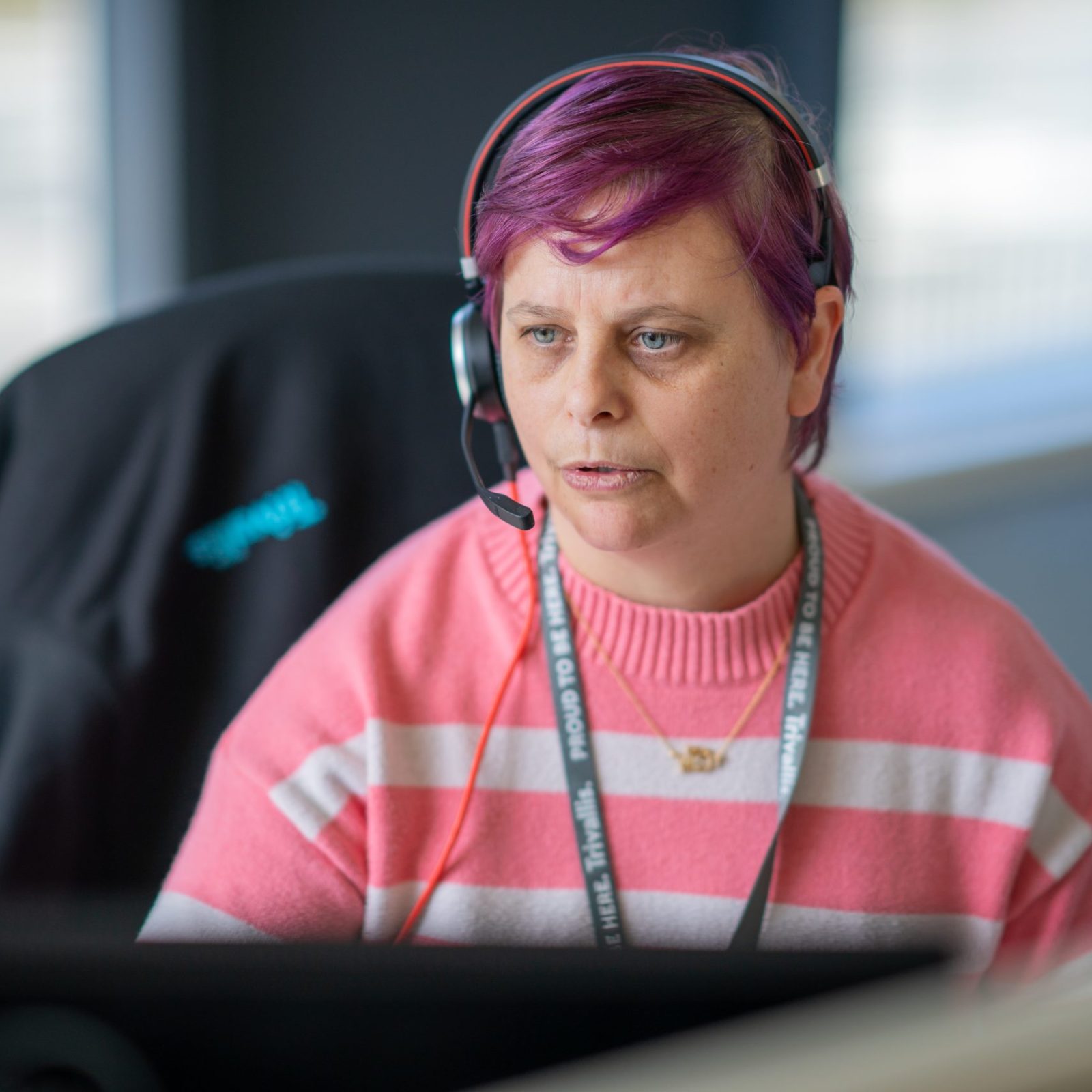 Trivallis Housing Landlord Wales A woman with purple hair wearing a headset sits in front of a computer monitor in an office setting. she is wearing a pink and white striped sweater and has a focused expression.