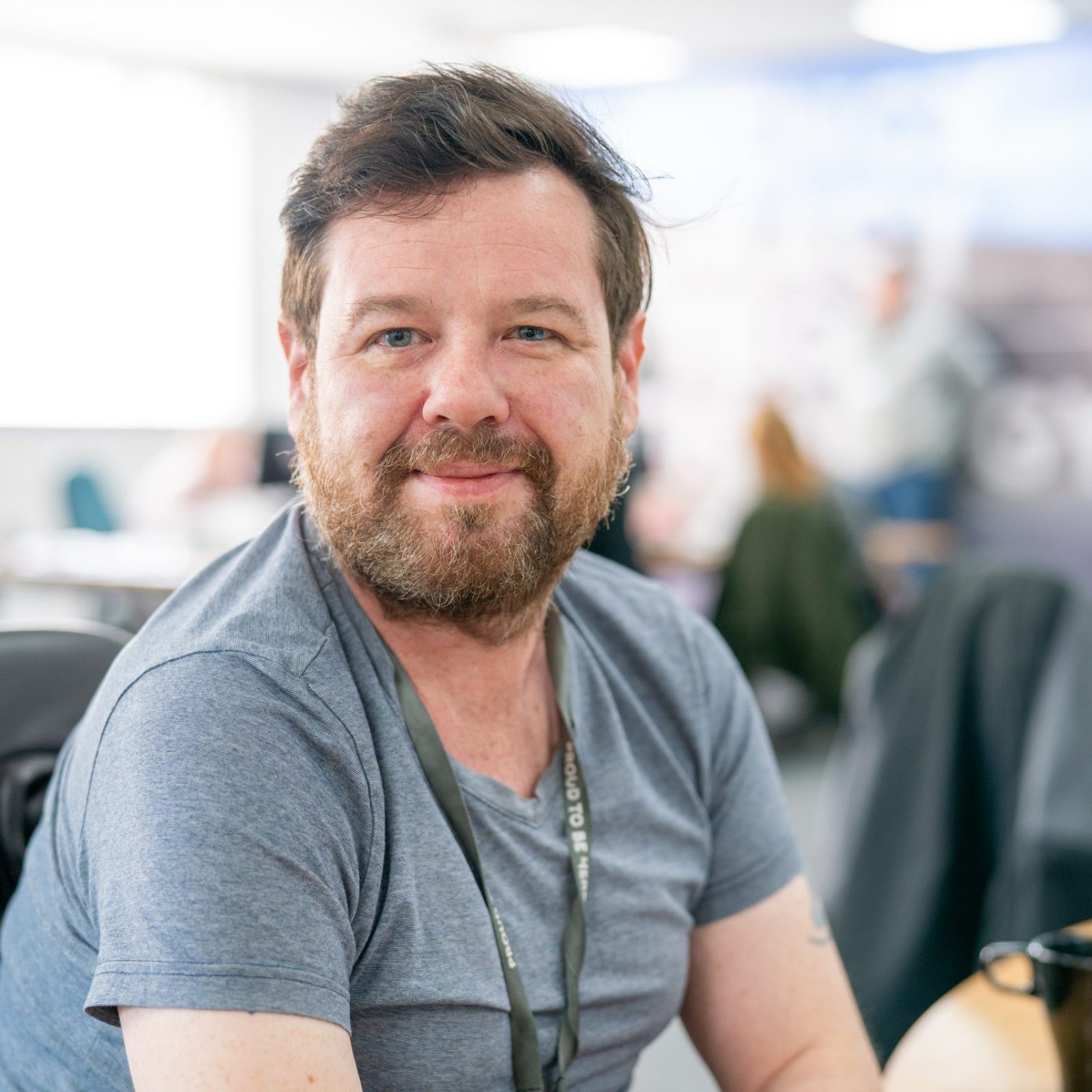 Trivallis Housing Landlord Wales A smiling man with facial hair sits at a desk in a busy office environment, wearing a gray t-shirt and a lanyard. he seems relaxed, looking directly at the camera with office colleagues blurred in the background. He is a money advice officer who can help with money.