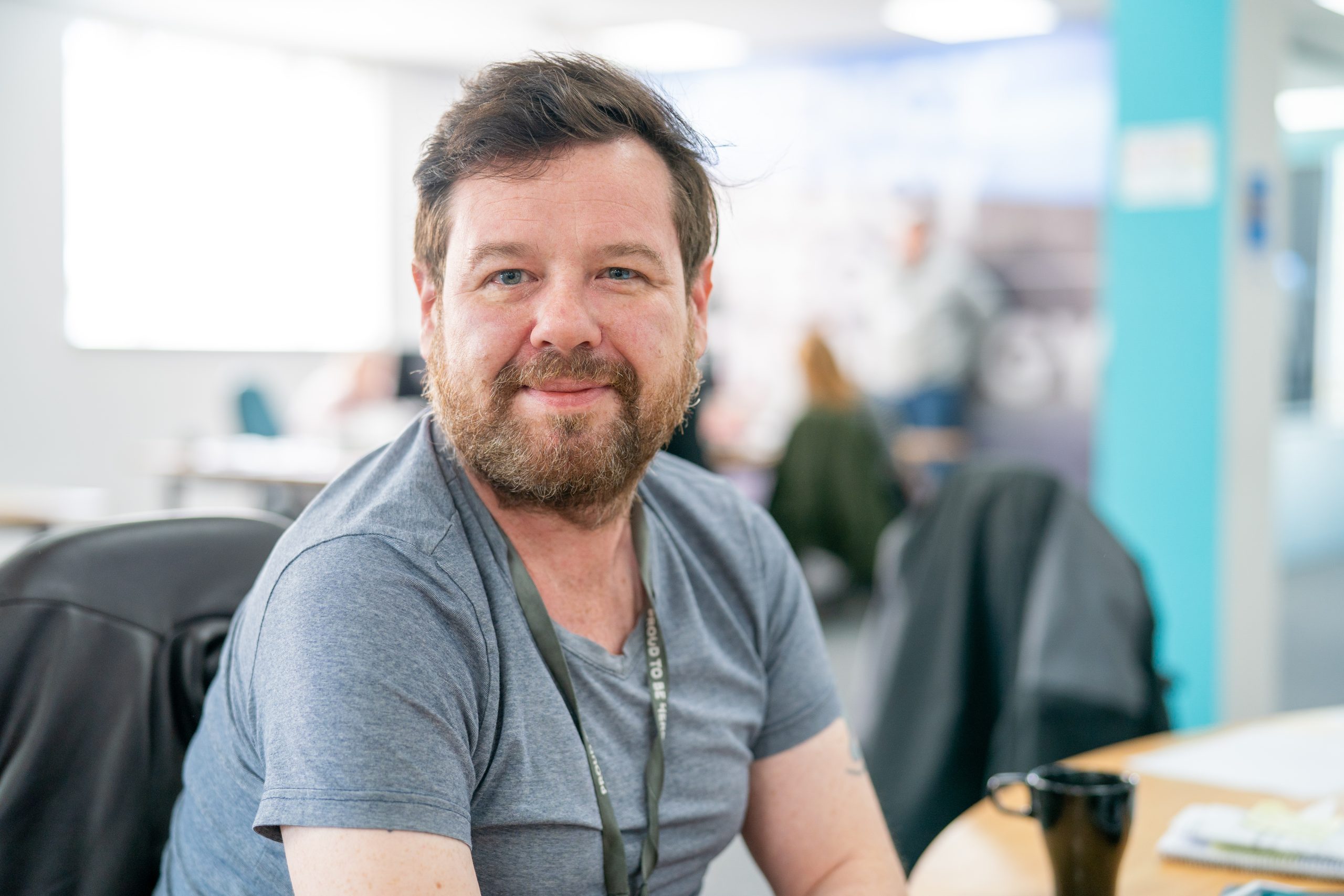 Trivallis Housing Landlord Wales A smiling man with facial hair sits at a desk in a busy office environment, wearing a gray t-shirt and a lanyard. he seems relaxed, looking directly at the camera with office colleagues blurred in the background. He is a money advice officer who can help with money.