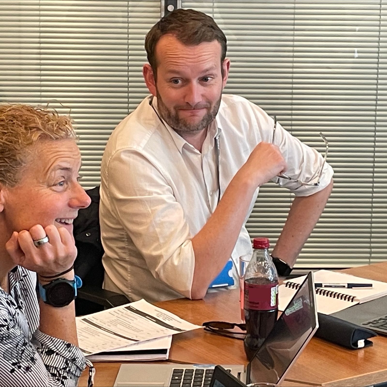 Trivallis Housing Landlord Wales Two professionals, a man and a woman, in an office meeting room. The woman is glancing sideways with a smile, and the man is looking at the camera with an elbow on the chair back. A bottle and laptops are visible.