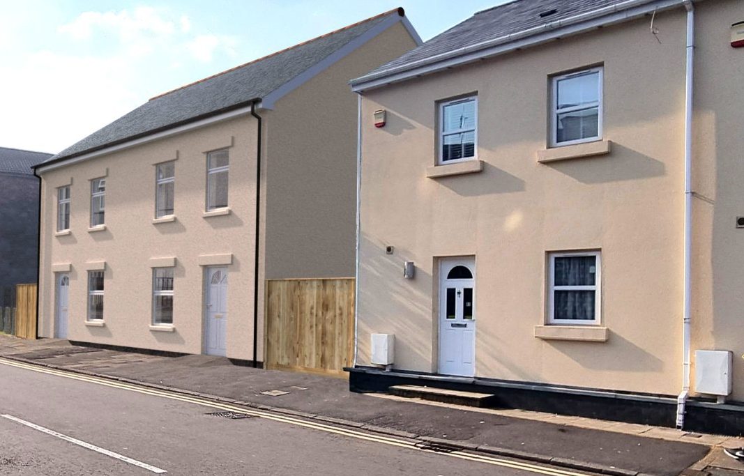 Trivallis Housing Landlord Wales A man in white walks past new light-colored townhouses on a sunny street with cars parked along the curb, with other residential buildings and a clear blue sky in the background. Safety checks keep these homes safe from hazards.