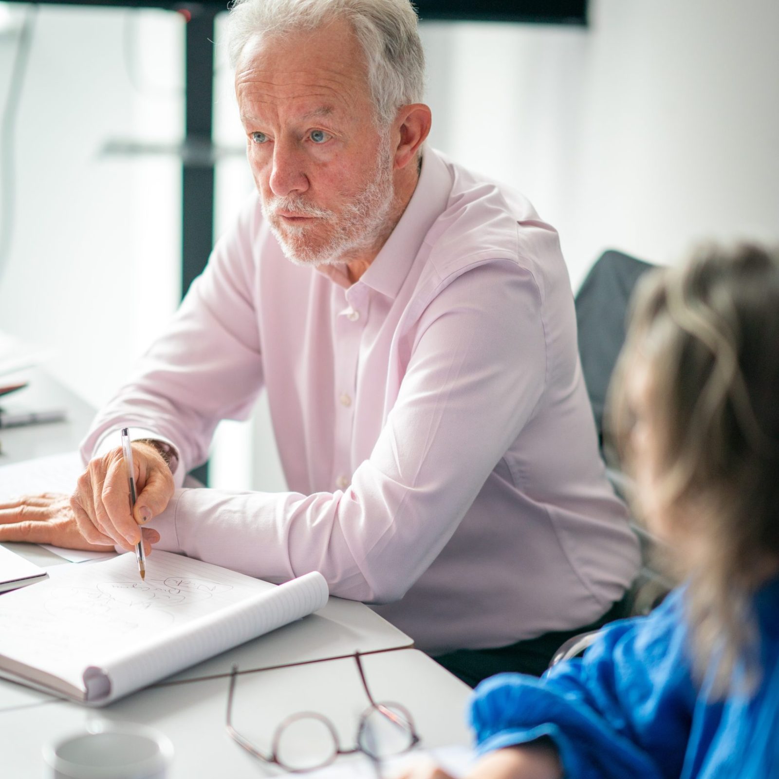 Trivallis Housing Landlord Wales An older man with white hair and a beard, dressed in a light pink shirt, is seated at a table with an open notebook, holding a pen as if discussing something. A person in a blue sweater, whose face is out of focus, is also at the table with notes.