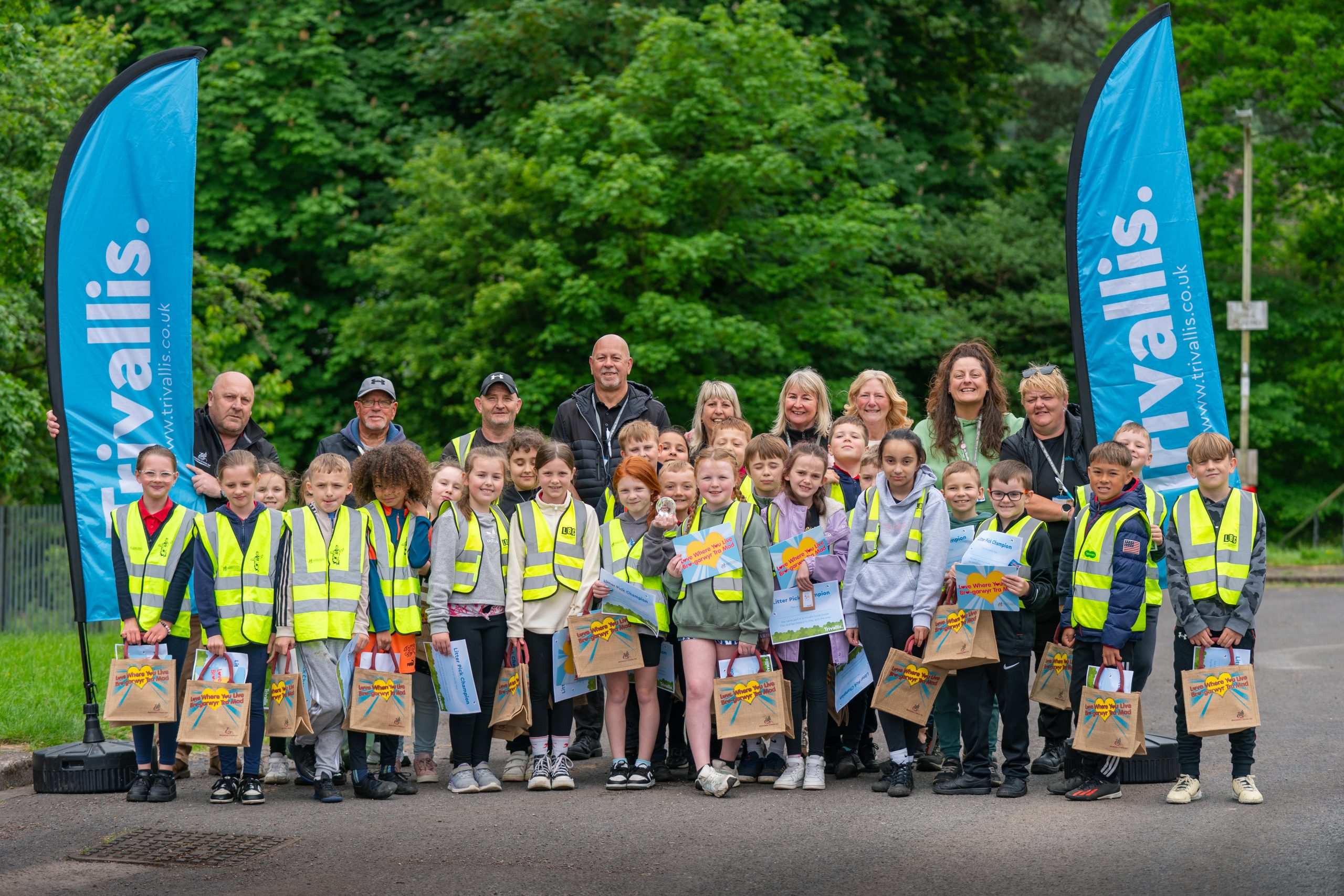 Trivallis Housing Landlord Wales A group photo shows children in high-visibility vests standing in front of adults and two blue flags with the word "Kivallis" on them. Each child holds a goodie bag. The background features green trees, indicating the photo was taken outdoors.