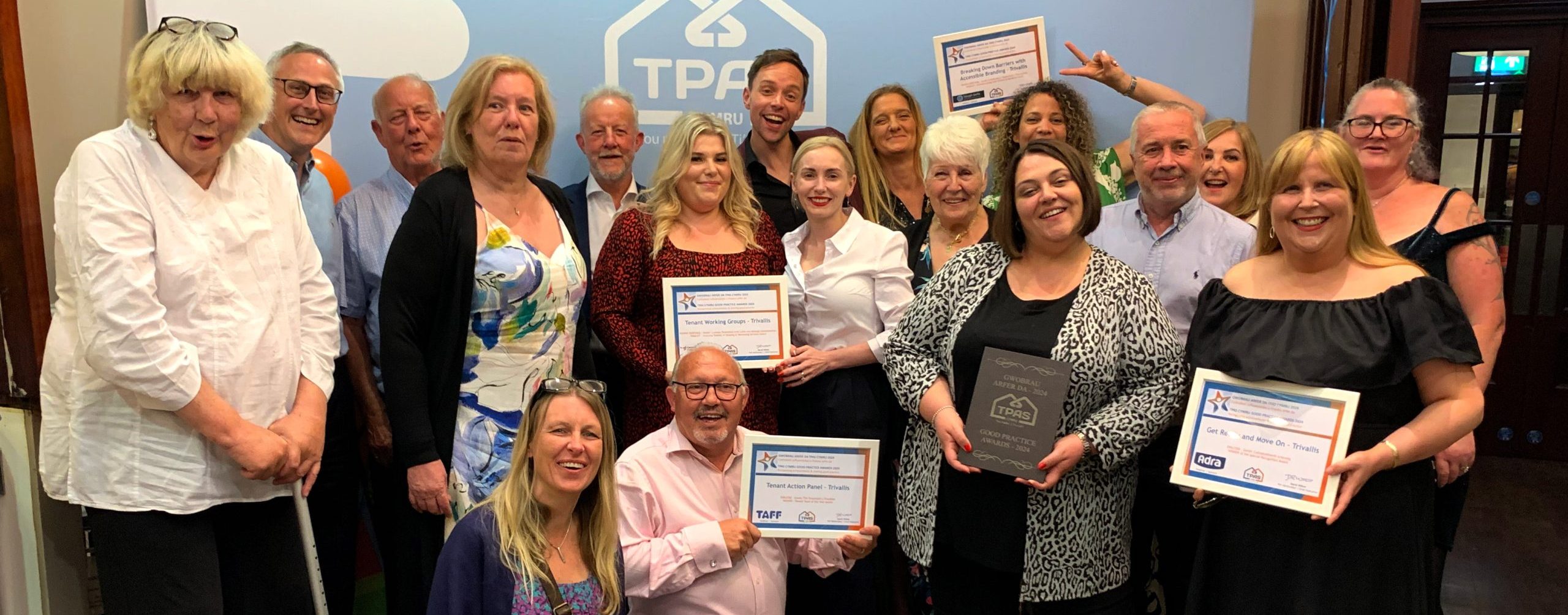 Trivallis Housing Landlord Wales A group of people is posing for a photo indoors. Some individuals are holding certificates and an award plaque. They are standing in front of a backdrop with the TPA logo. The group is casually dressed, and they appear to be celebrating an achievement.