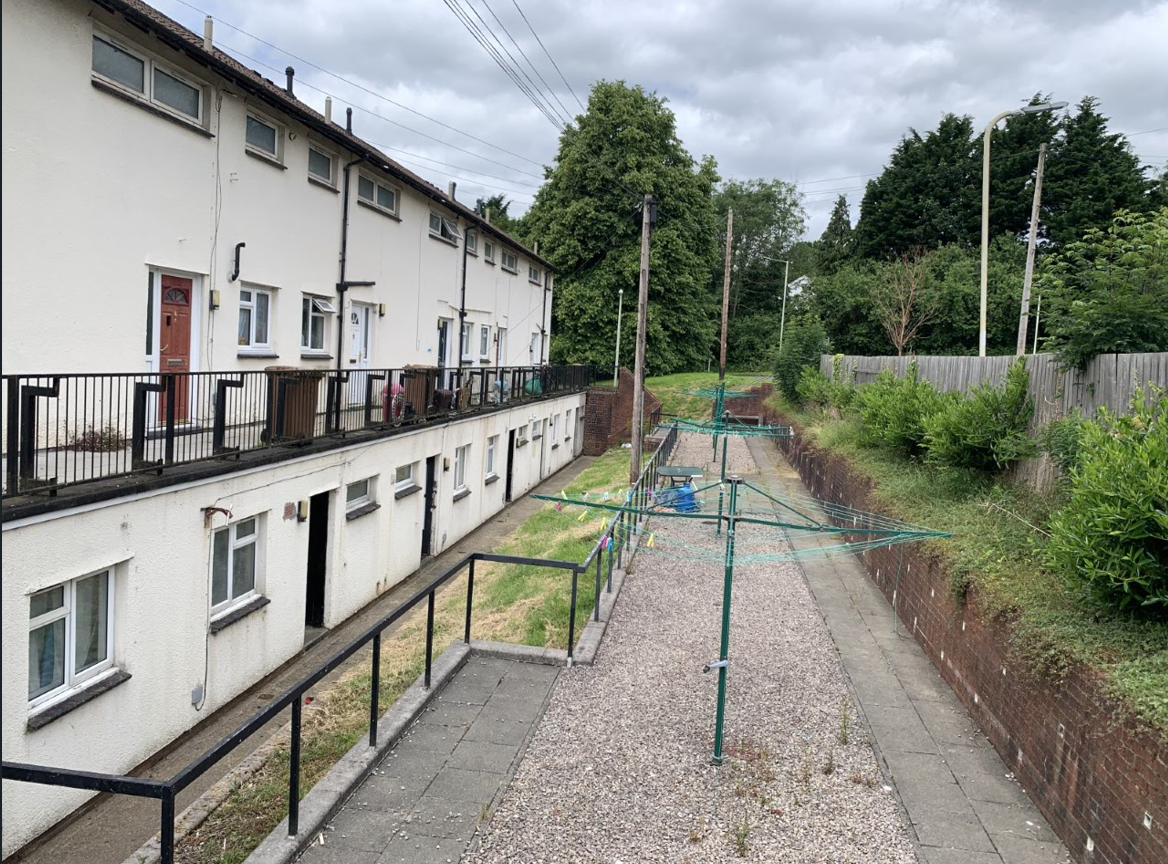 Trivallis Housing Landlord Wales A view of a backyard area behind a row of white apartment buildings. The area includes a gravel pathway, a green rotary clothesline, and is surrounded by grass and trees. There are concrete steps leading down from the buildings, and a wooden fence on one side.