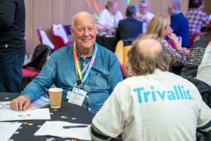 Trivallis Housing Landlord Wales Two people are sitting at a table during a social event. One man with a lanyard smiles, while the other wears a shirt with "Trivallis" on the back. Papers and a cup are on the table, and others are interacting in the background.