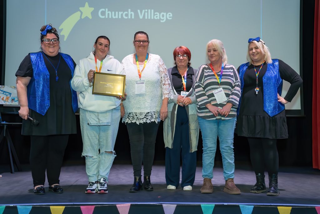 Trivallis Housing Landlord Wales A group of six people stand on a stage. One person holds a plaque. They are in front of a screen displaying the words "Church Village." Multicolored bunting decorates the front of the stage.