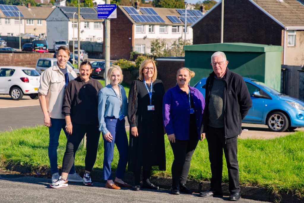 Trivallis Housing Landlord Wales A group of six people stand together outdoors on a sunny day. They are smiling near a grass area, with houses and parked cars in the background. A blue sign is visible behind them.