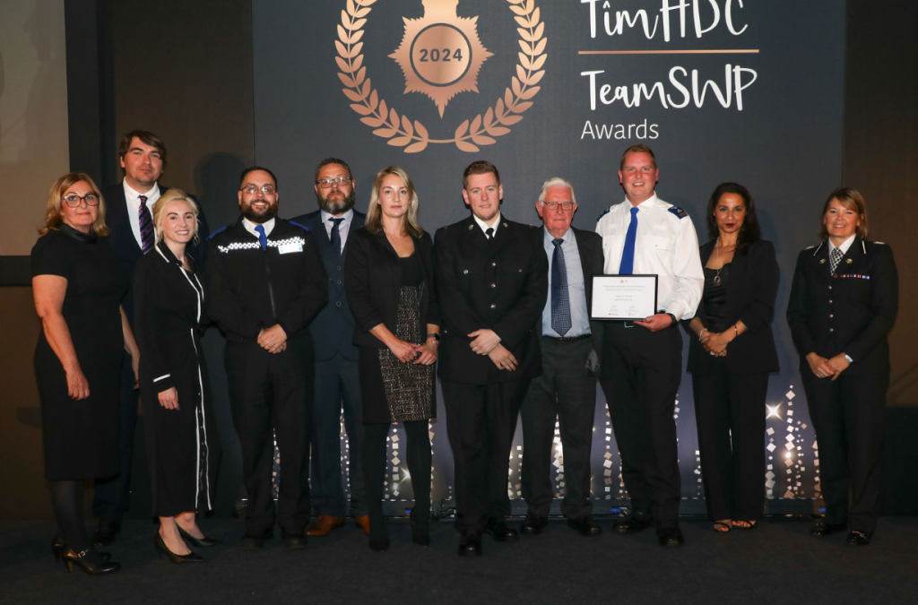 Trivallis Housing Landlord Wales A group of eleven people, including individuals in various uniforms and formal attire, stand on stage at an awards event. One individual holds a certificate. A large backdrop displays "Team HDC Team SWP Awards 2024" with decorative elements.