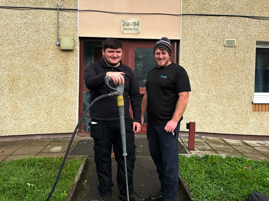 Trivallis Housing Landlord Wales Two men dressed in work clothes stand outside a building. One man holds a power tool connected to a hose. The building has a sign reading "29-34 Guardian Place." There's a grass lawn and a paved path in front of them.