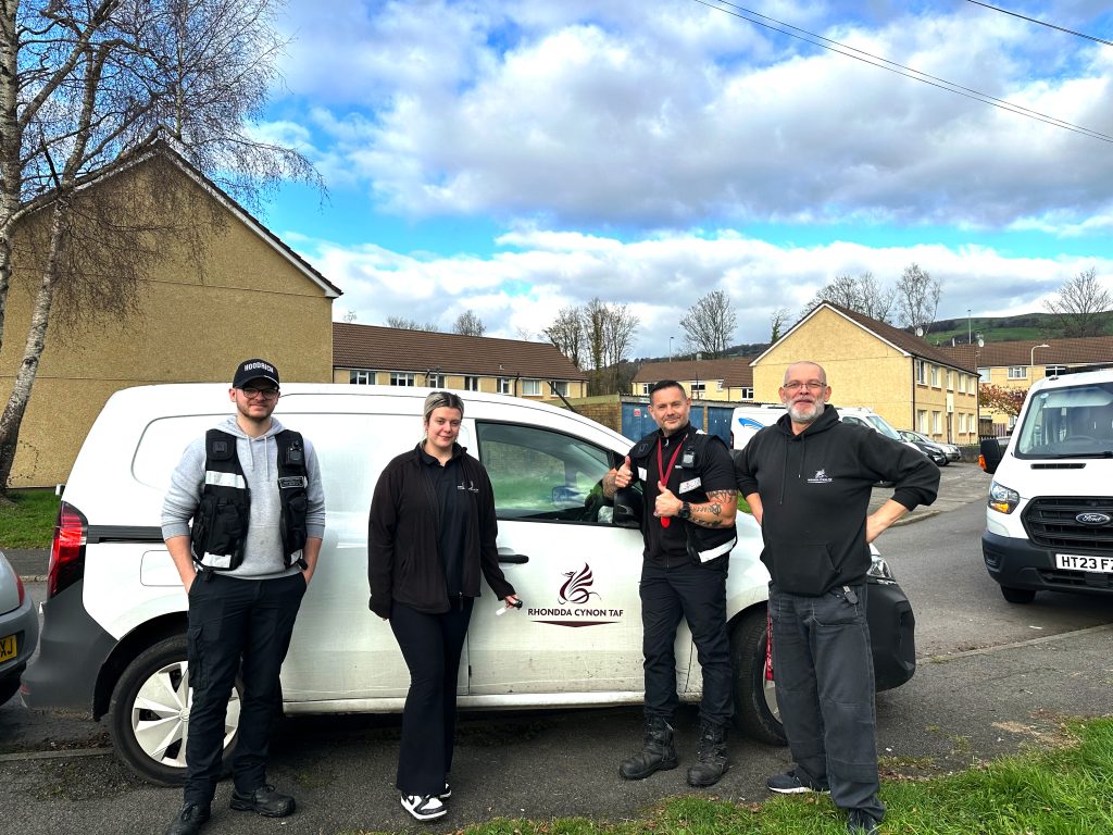 Trivallis Housing Landlord Wales Four people stand in front of a white van parked on a residential street. The van has a logo on the door. Two people give thumbs-up gestures. The background shows houses, a tree, and cloudy skies.