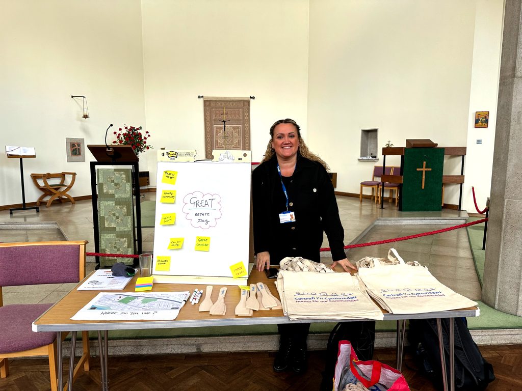 Trivallis Housing Landlord Wales A person stands behind a table inside a church. The table displays a board with sticky notes, tote bags, a map, and wooden utensils. Church furnishings are seen in the background.