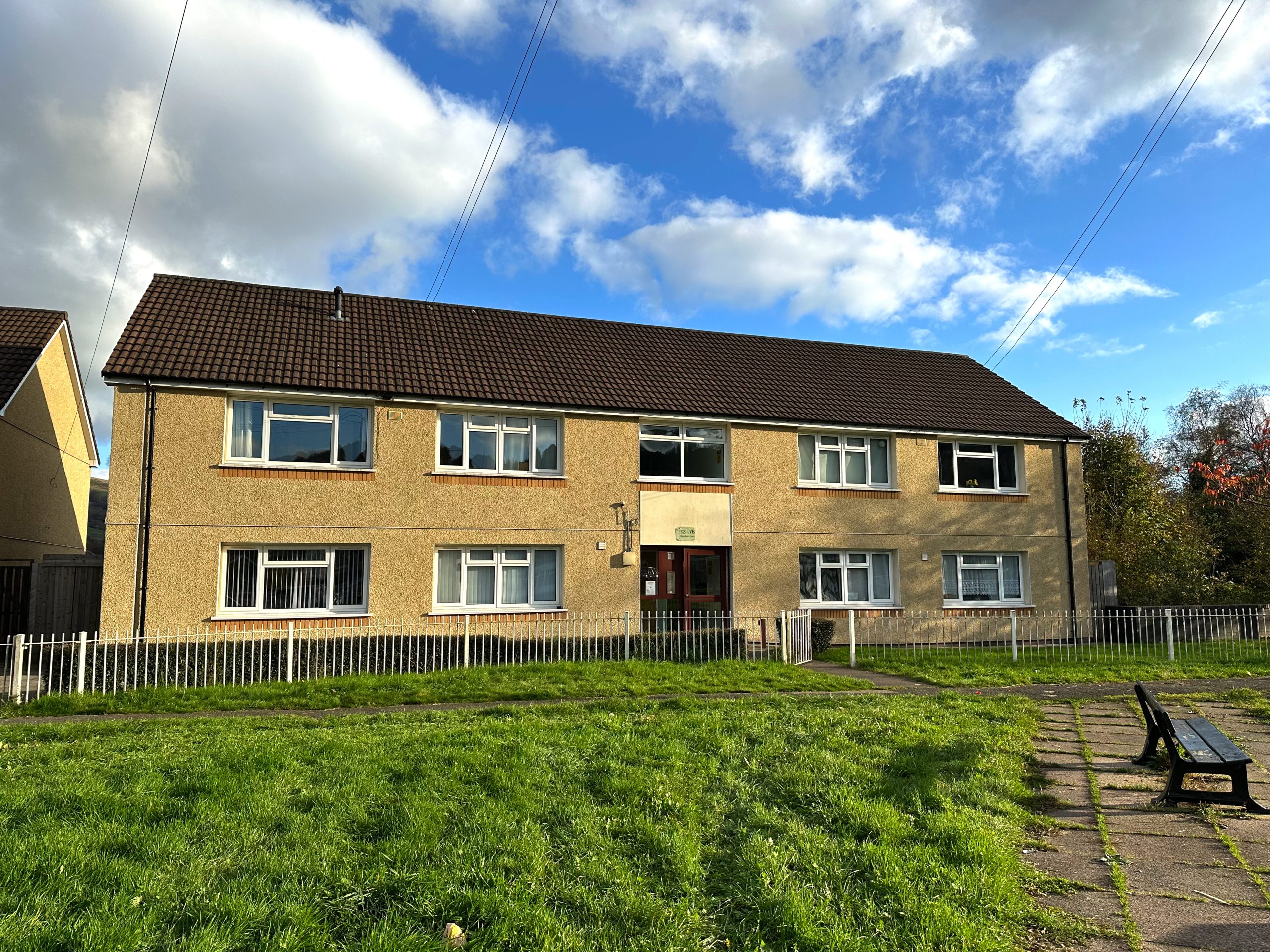 Trivallis Housing Landlord Wales A two-story apartment building with a brown tiled roof and beige exterior. It has multiple windows on both floors. The front yard contains a grassy area, a concrete path, and a bench, all bordered by a metal fence. The sky is partly cloudy.