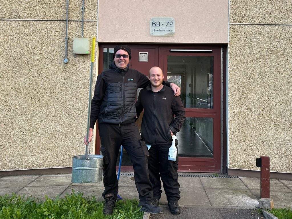 Trivallis Housing Landlord Wales Two men stand outside a building entrance labeled 69-72 with the sign "Glenshiel Place" above the door. One man wears sunglasses and has his arm around the other. They are dressed in work clothes and holding cleaning equipment.