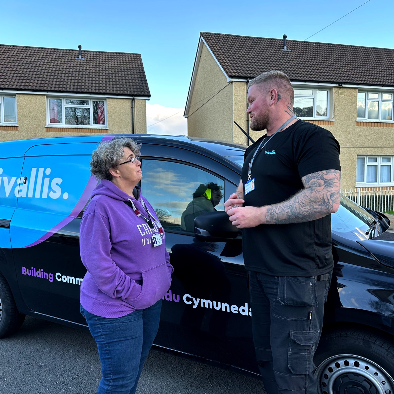 Trivallis Housing Landlord Wales A woman in a purple hoodie talks to a man in a black shirt standing next to a blue and black Trivallis van. They are in a residential area with houses in the background.