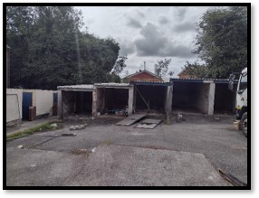 Trivallis Housing Landlord Wales An outdoor scene showing a set of four empty, open garages with wear and broken doors. A truck is parked to the right. Trees and cloudy sky are in the background. Debris is scattered on the ground.