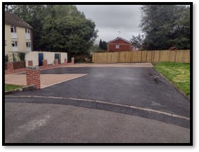 Trivallis Housing Landlord Wales Driveway entrance with a paved section on the left and a tarmac surface. Brick wall and several small storage units are visible. Residential buildings are in the background, with trees and a wooden fence lining the area. Cloudy sky overhead.