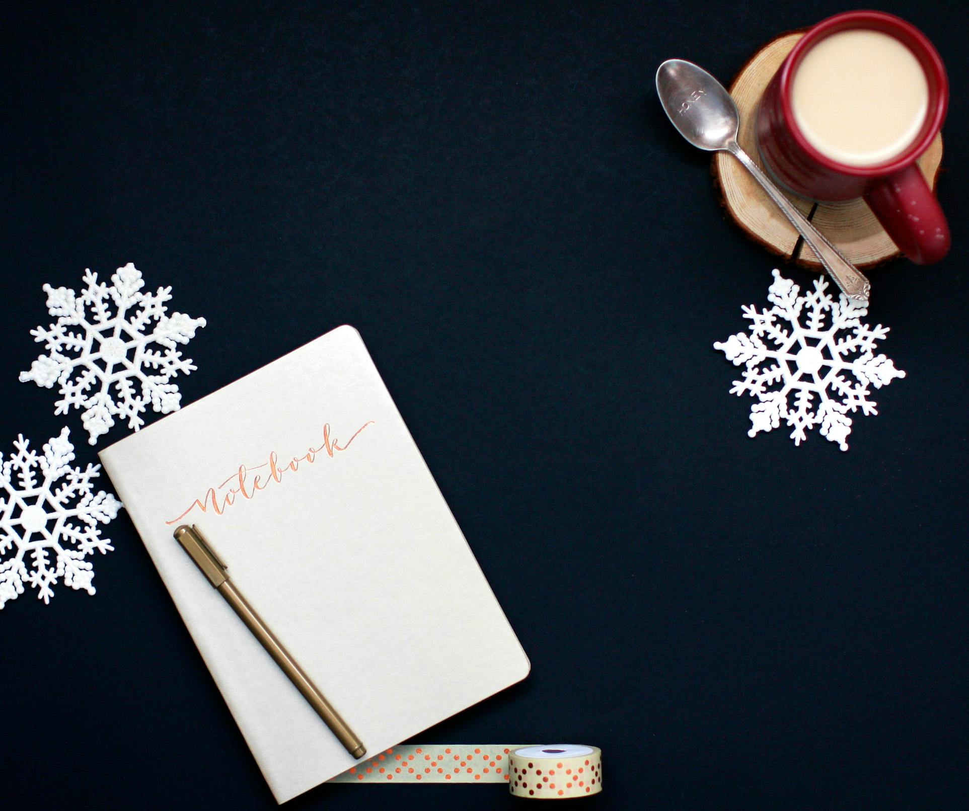 Trivallis Housing Landlord Wales A notebook with a pen rests on a dark surface, accompanied by two white snowflake decorations. A red mug with a spoon on a wooden coaster holds a light-colored beverage. A roll of washi tape is nearby.