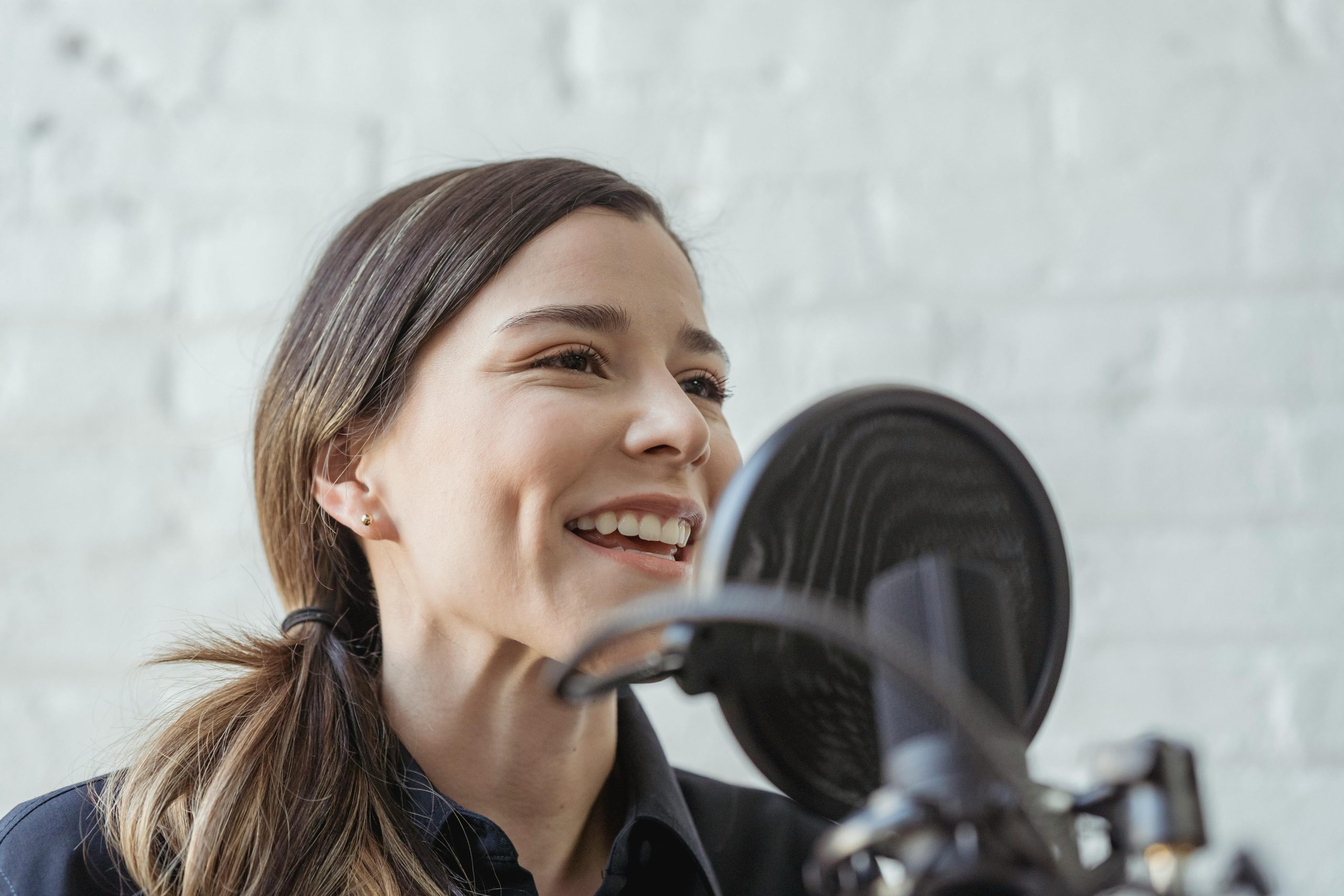 Trivallis Housing Landlord Wales A person with long hair smiles while speaking into a microphone, protected by a pop filter, with a white brick wall in the background for a podcast.