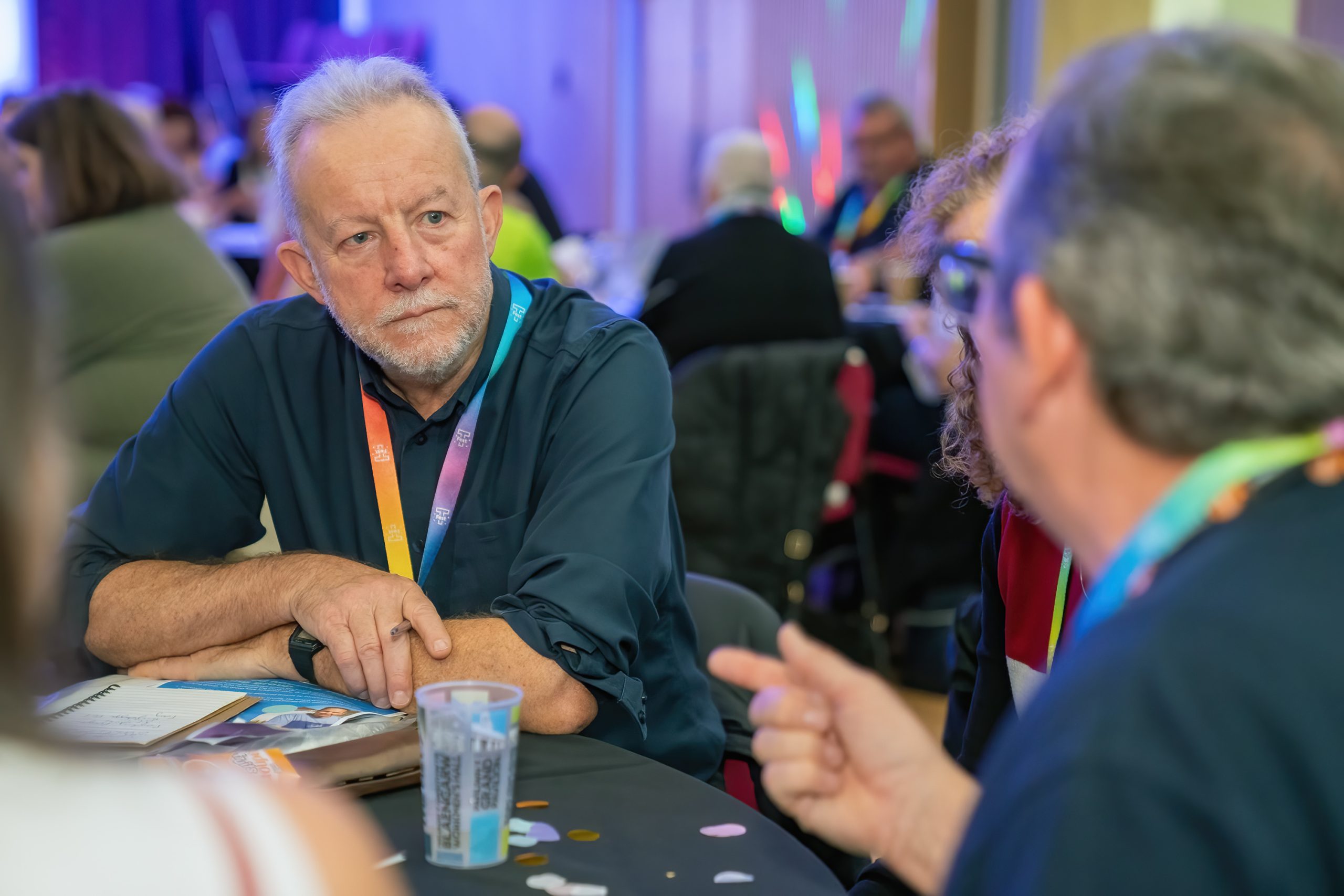 Trivallis Housing Landlord Wales A group of people sitting at a round table engaged in conversation. One man with gray hair and a blue shirt is listening intently to another person speaking. The table has cups, papers, and some colorful confetti on it.