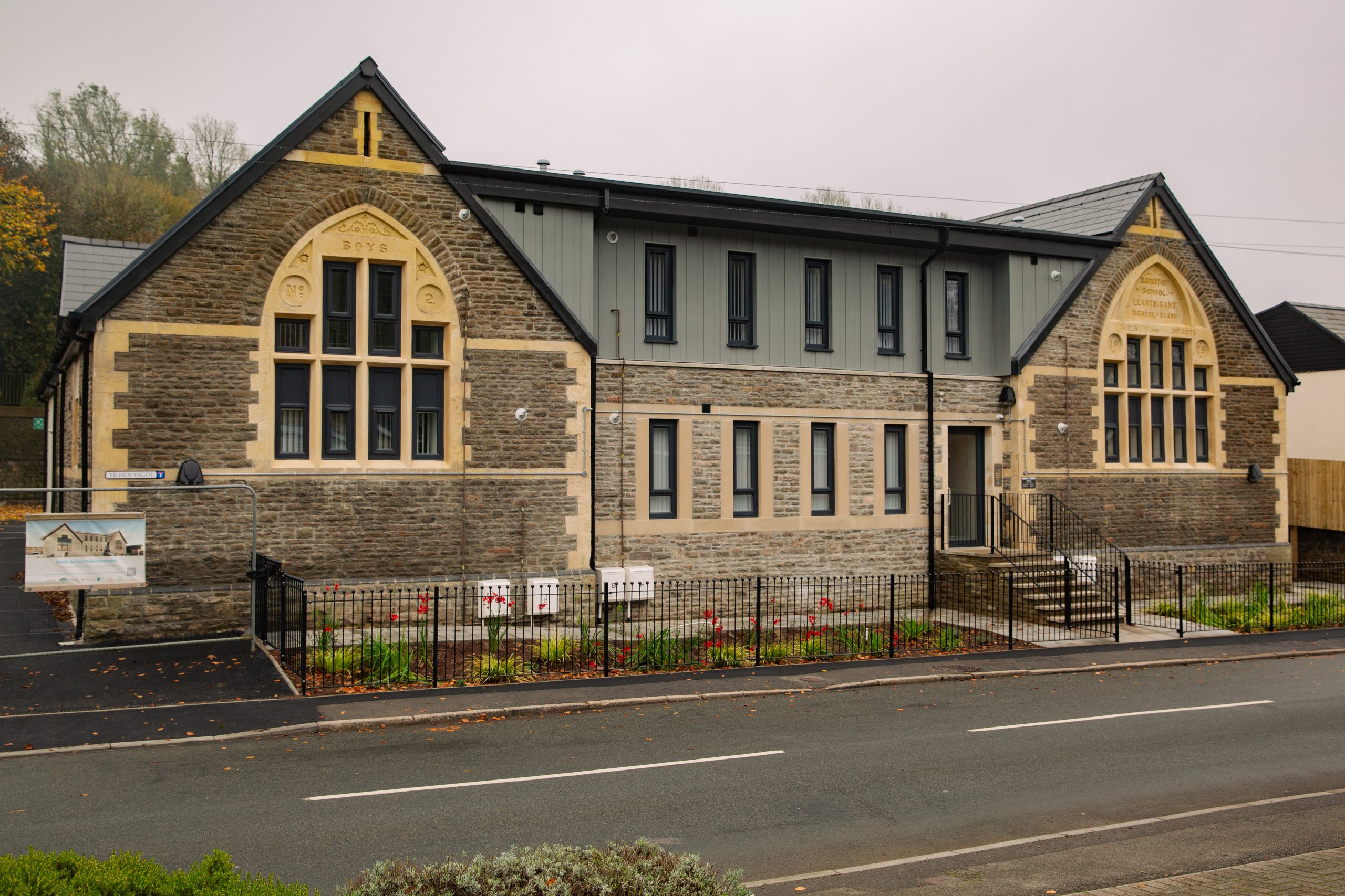 Trivallis Housing Landlord Wales A modernized building with a mix of stone and contemporary materials, featuring large windows and two prominent entrances. It has a landscaped front yard and is located along a road with trees in the background.