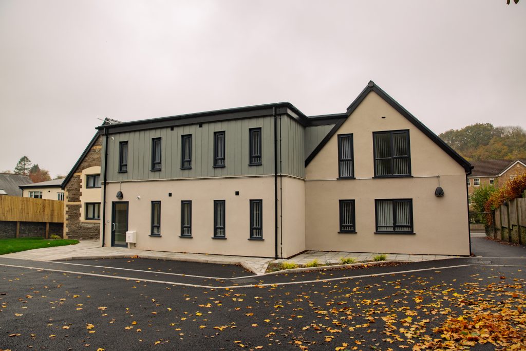 Trivallis Housing Landlord Wales A modern two-story building with a combination of cream and dark gray exterior. It has vertical windows and a sloping roof. The surrounding area features a paved road and scattered autumn leaves, with additional buildings and trees in the background.